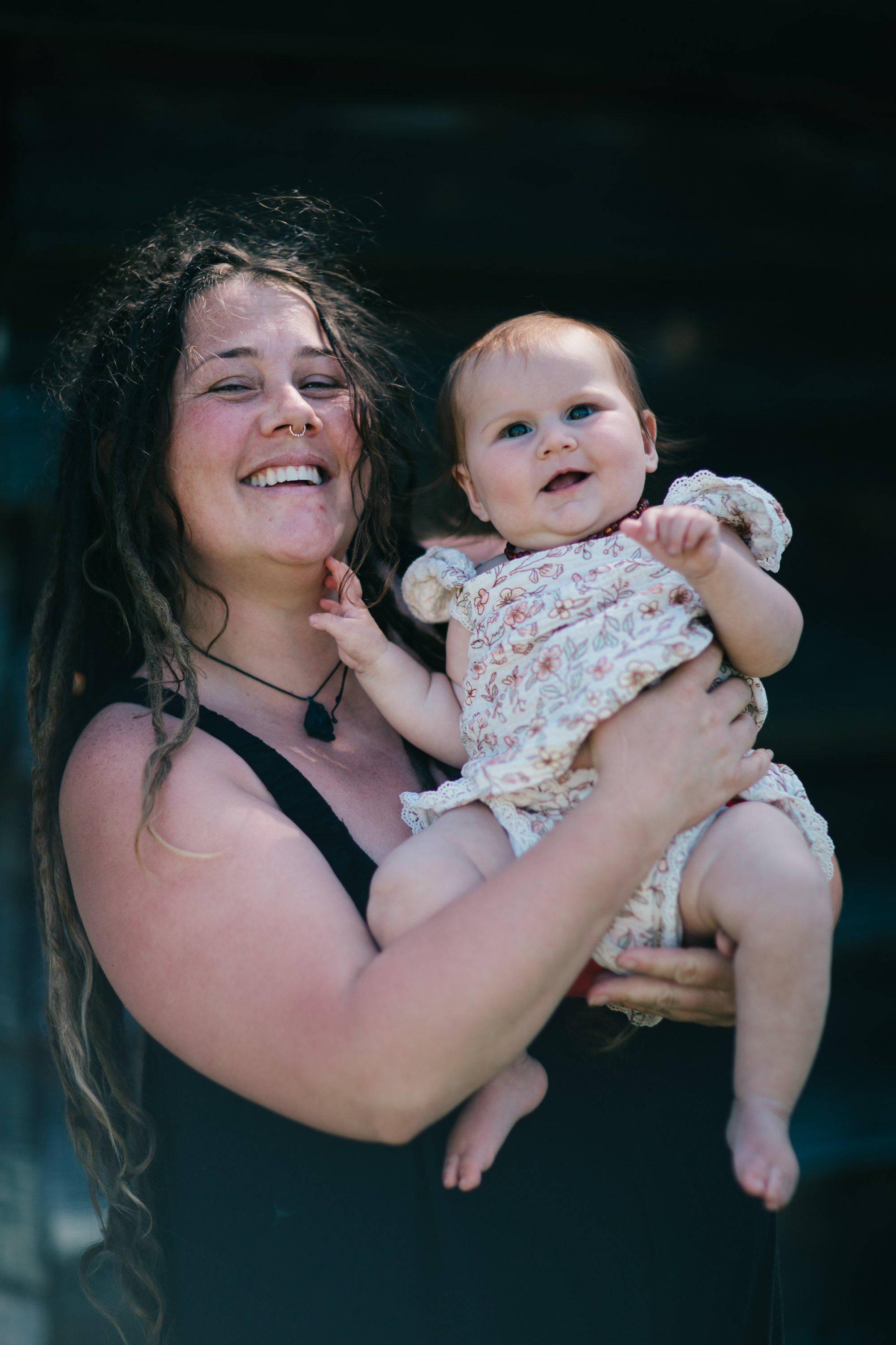 Woman holding smiling infant in floral dress, both joyfully gazing downward in natural light against dark background