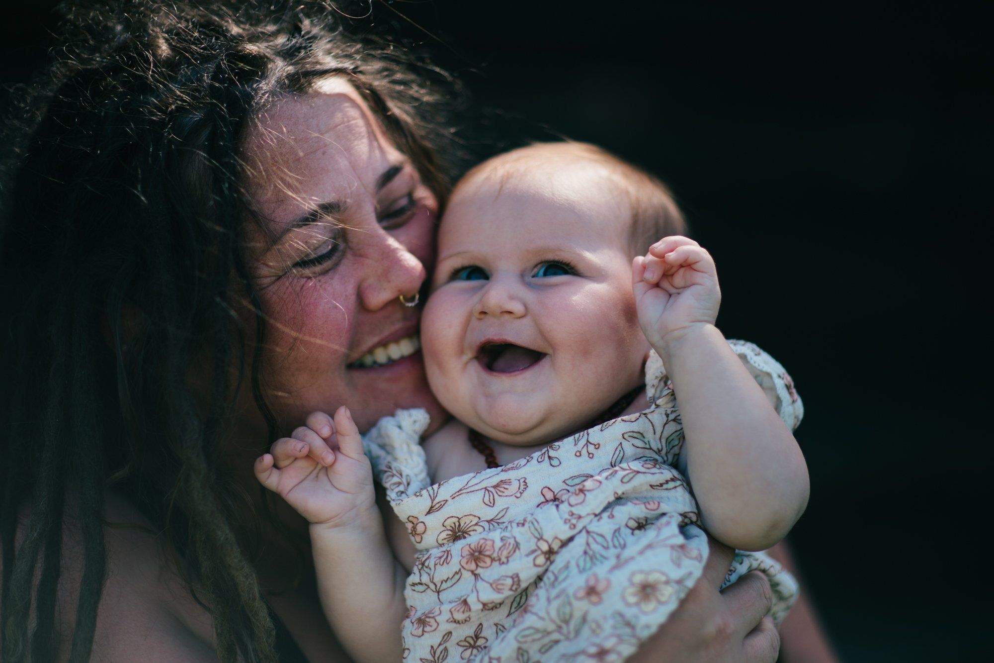 Woman with dark hair smiling warmly while holding a laughing baby in a patterned outfit against a dark background