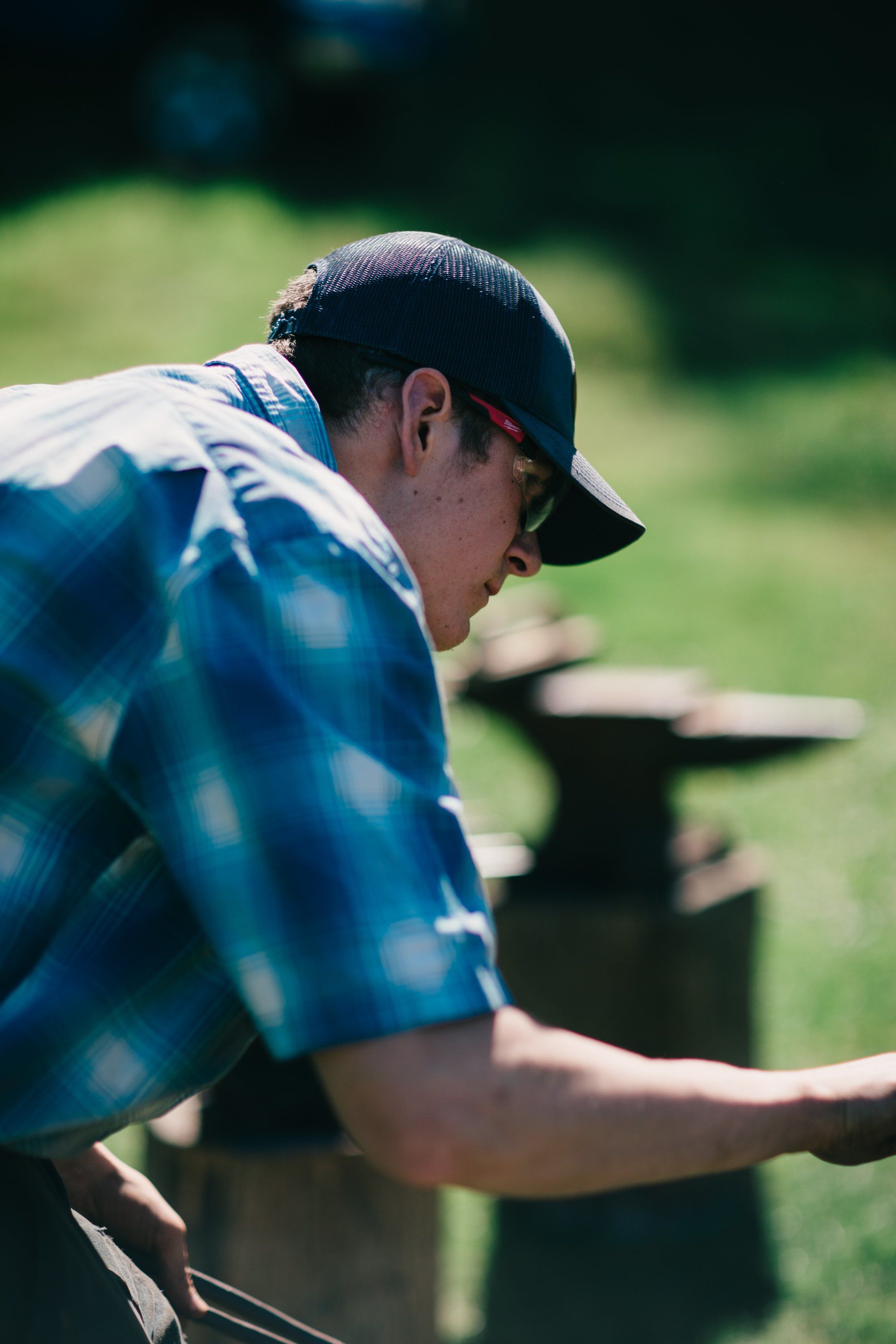 Man in blue plaid shirt and cap leans over wooden fence in sunlit pasture, focused on farm work