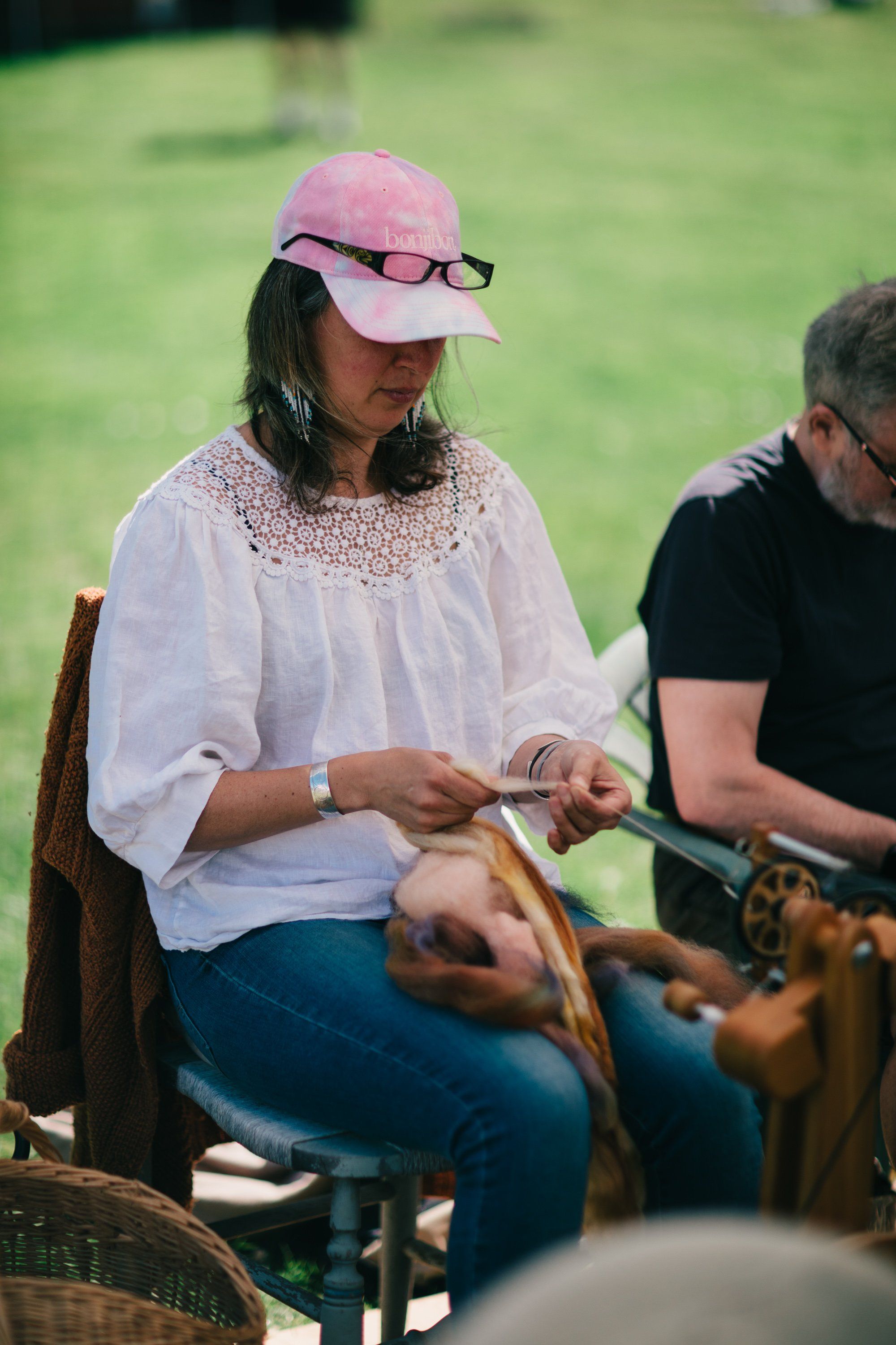 Woman in pink cap and white lace blouse works with rope outdoors, seated on a bench with green fields behind her