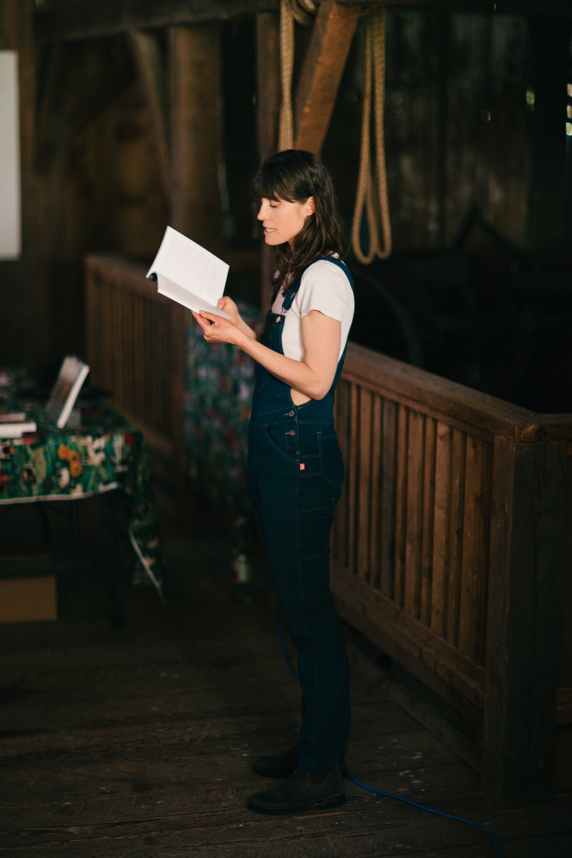 Woman in overalls holding papers stands in a rustic barn, warm light illuminating her against wooden beams and rope