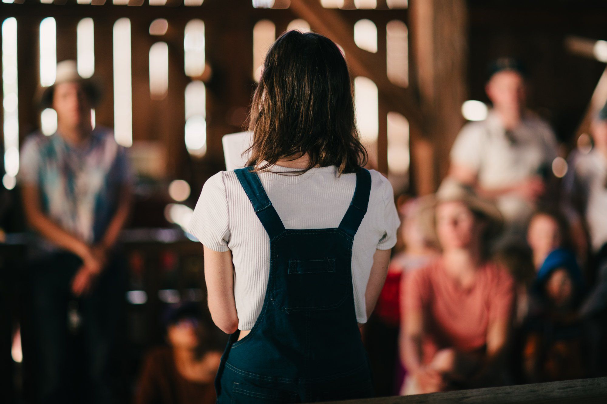 Young woman in overalls and white shirt stands facing away toward a gathered crowd in warm evening light at a farm venue