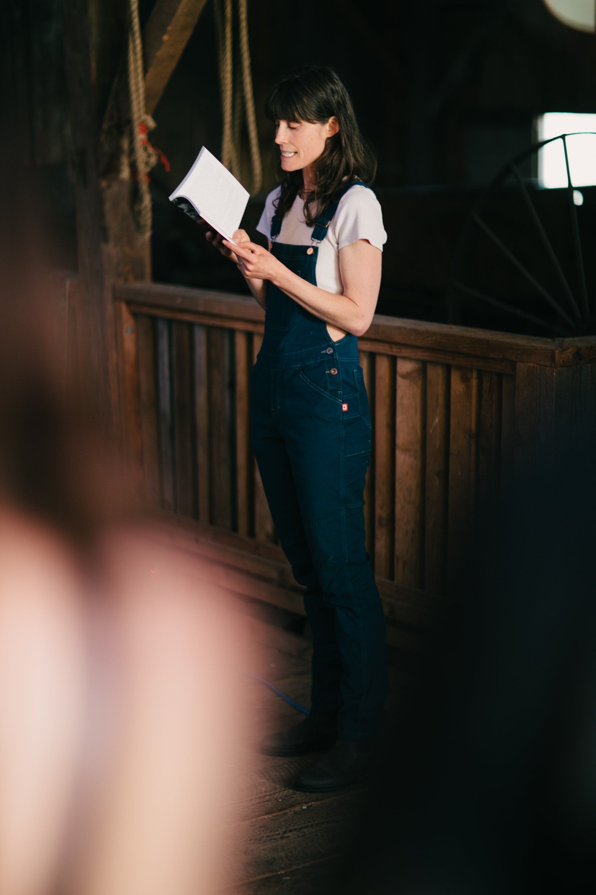 Woman holding script on wooden deck at dusk, warm stage lighting illuminating her against dark background