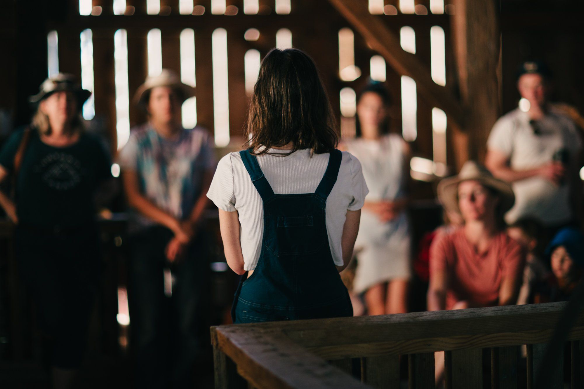 Woman in denim overalls stands facing a gathered crowd in a sunlit barn, backlit by wooden slats casting linear shadows