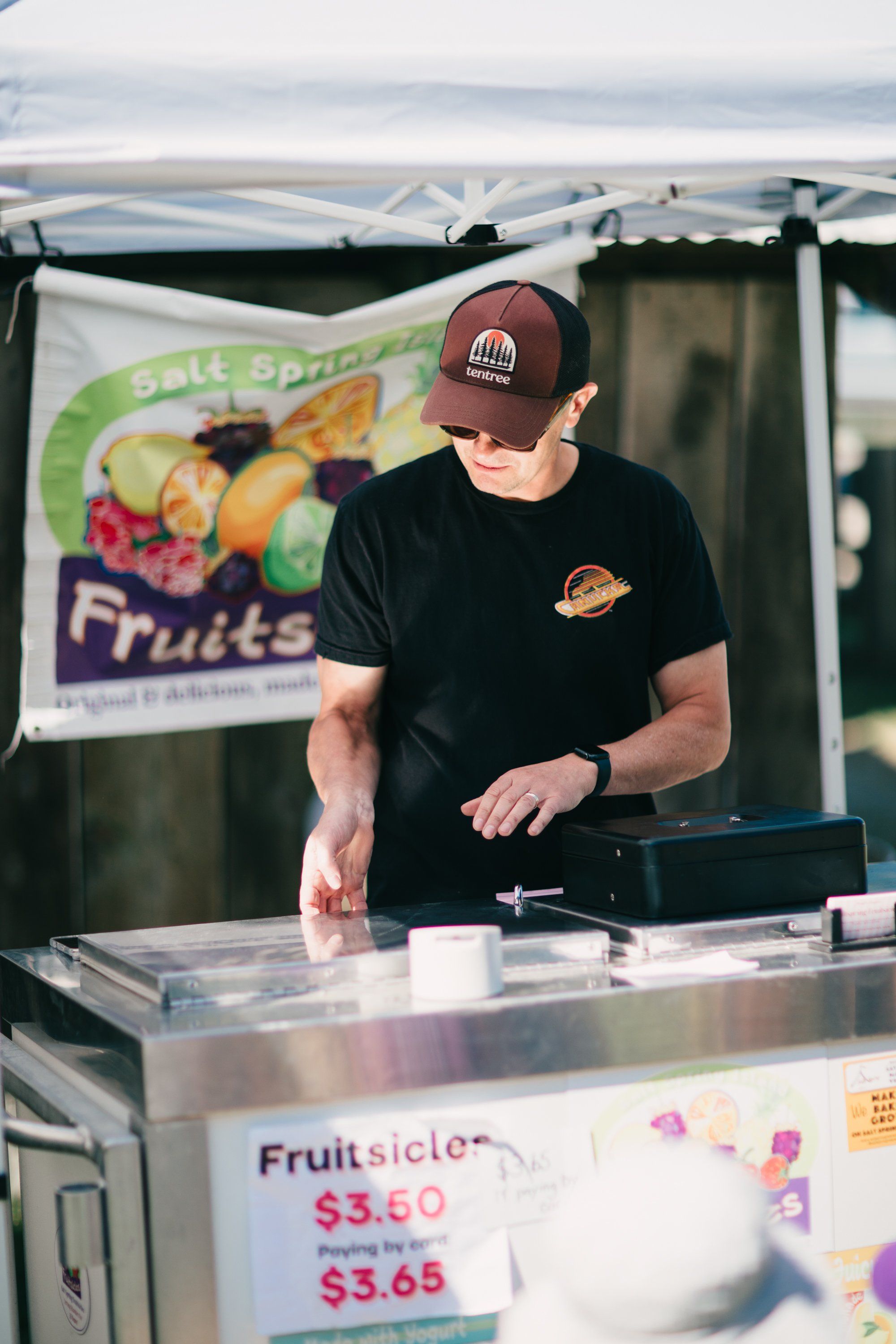Vendor in maroon cap and black shirt prepares fruit slices at farm stand under white tent canopy