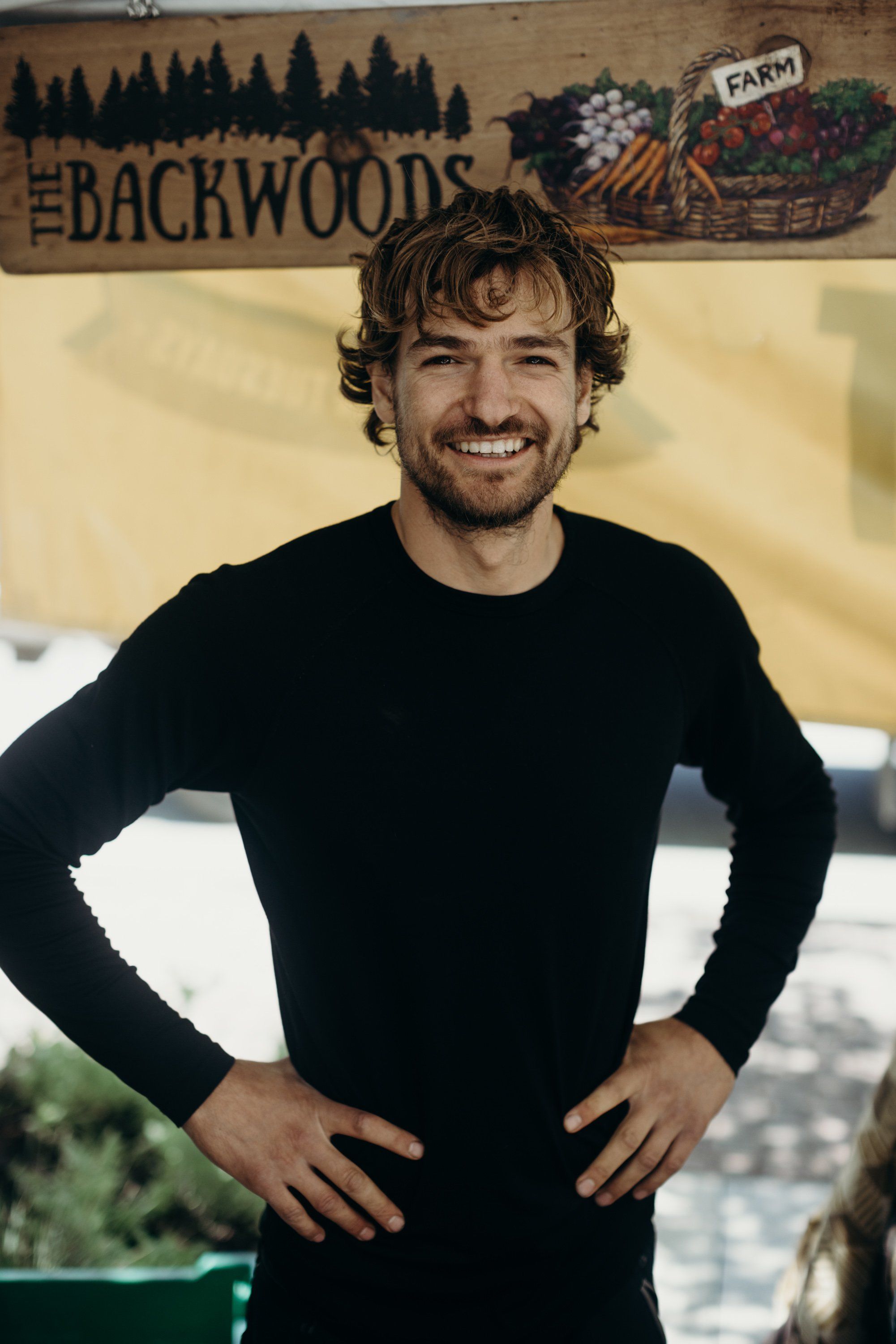 Smiling man with wavy brown hair in black long-sleeve shirt poses with hands on hips before rustic wooden backdrop