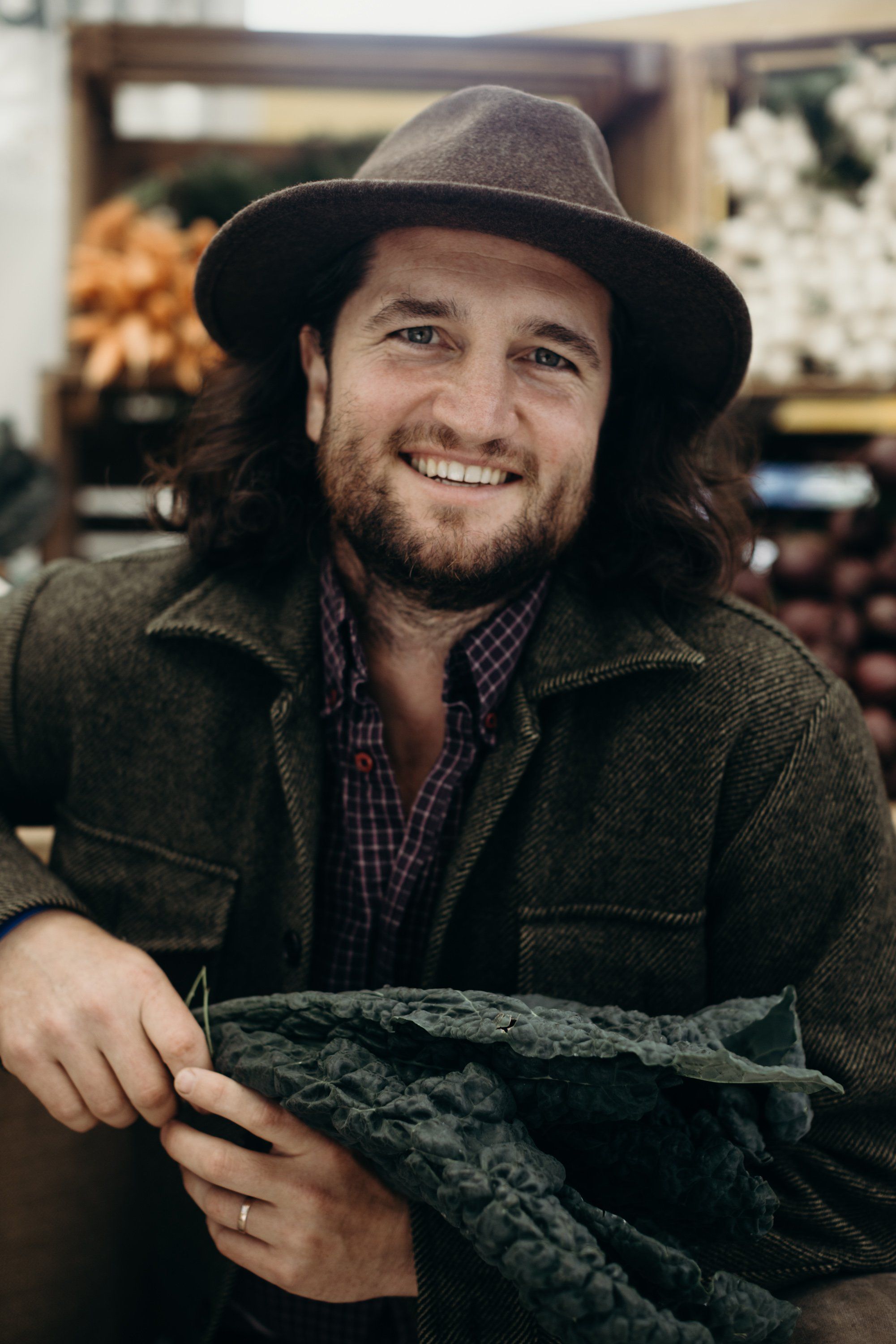 Smiling man in charcoal hat and olive jacket holds woven textile at indoor market, warm afternoon light