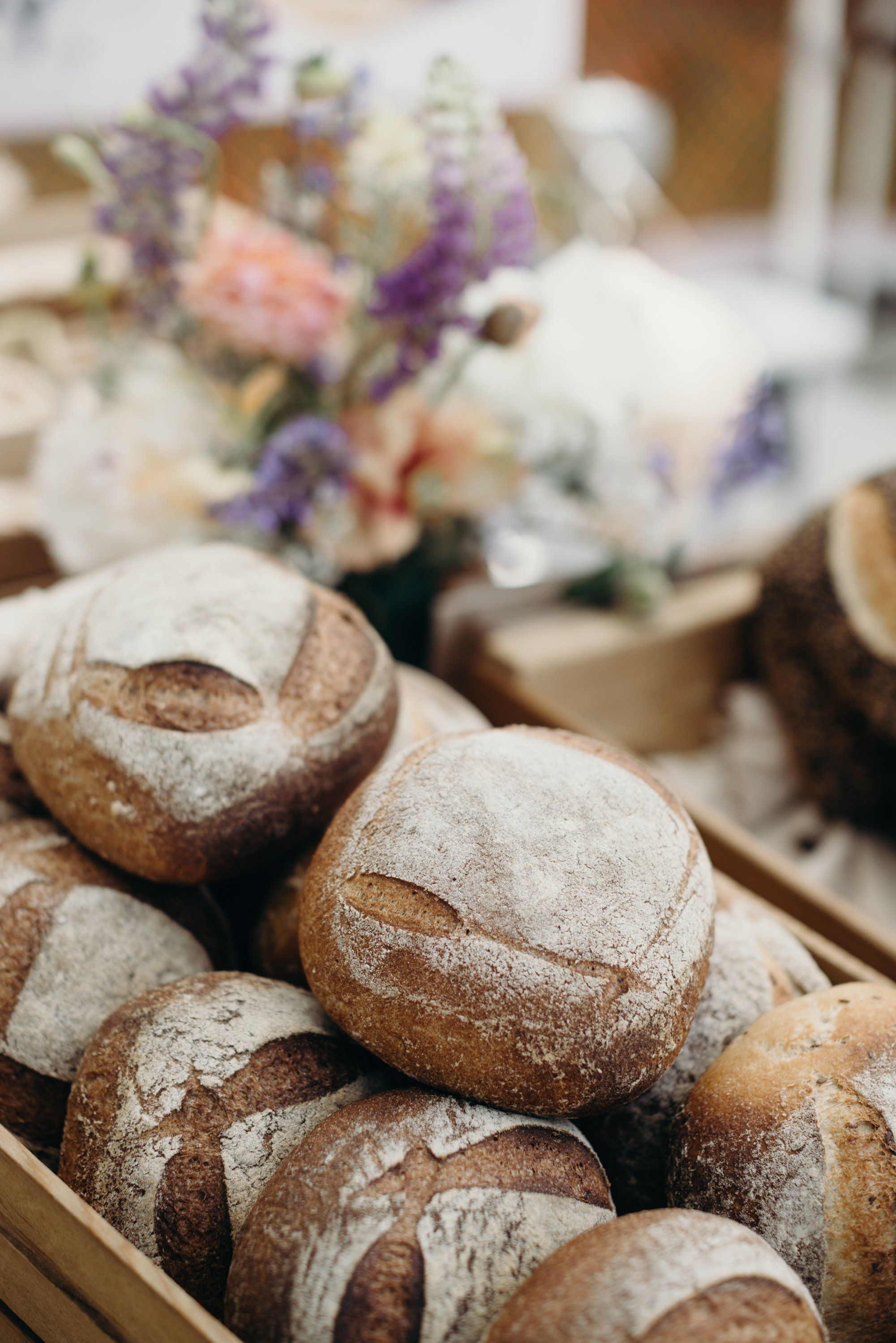 Crusty artisan bread rolls dusted with flour arranged in a wooden crate, with blurred flowers and market stalls behind