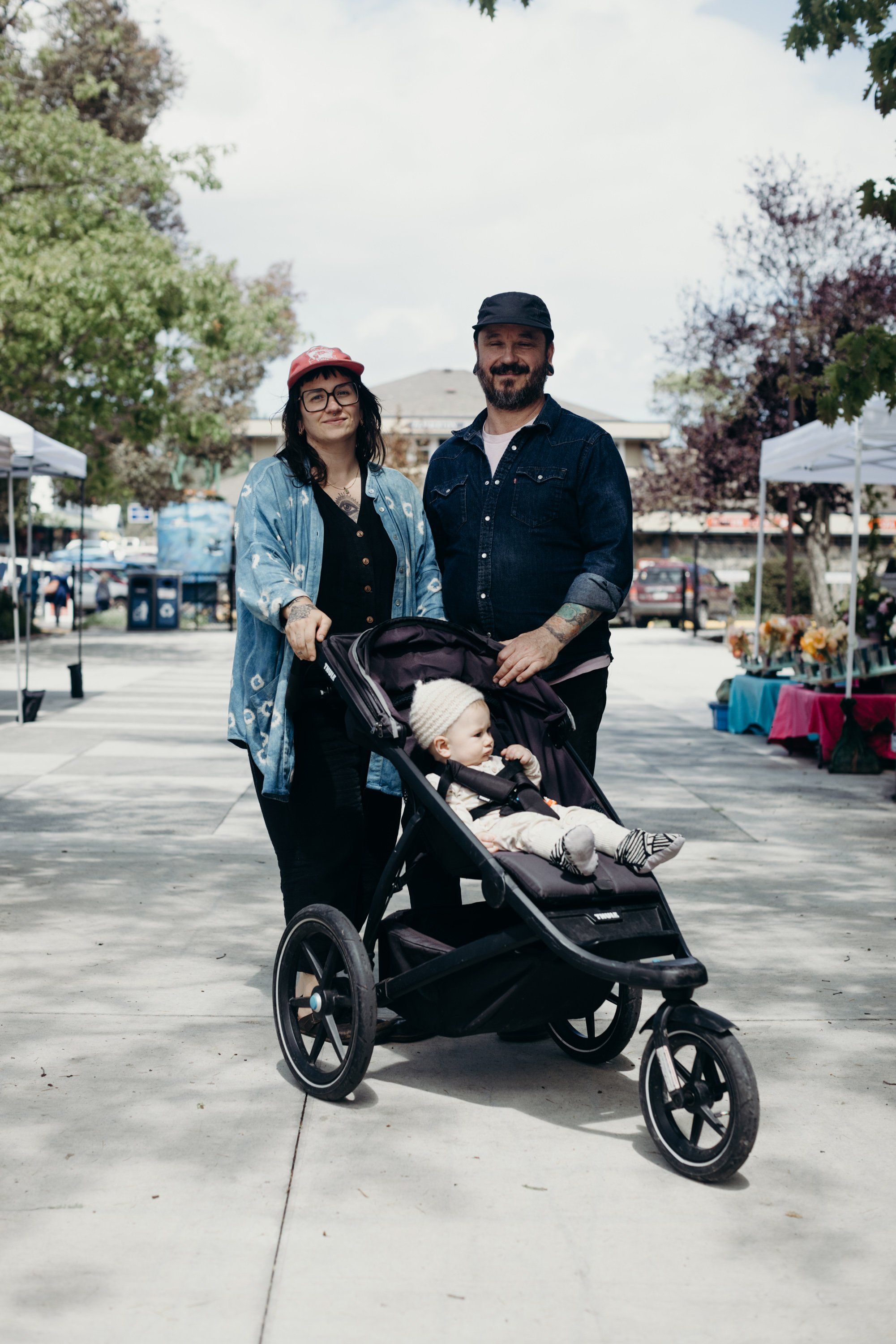 Couple with jogging stroller at farmers market on sunny day, tree-lined plaza setting