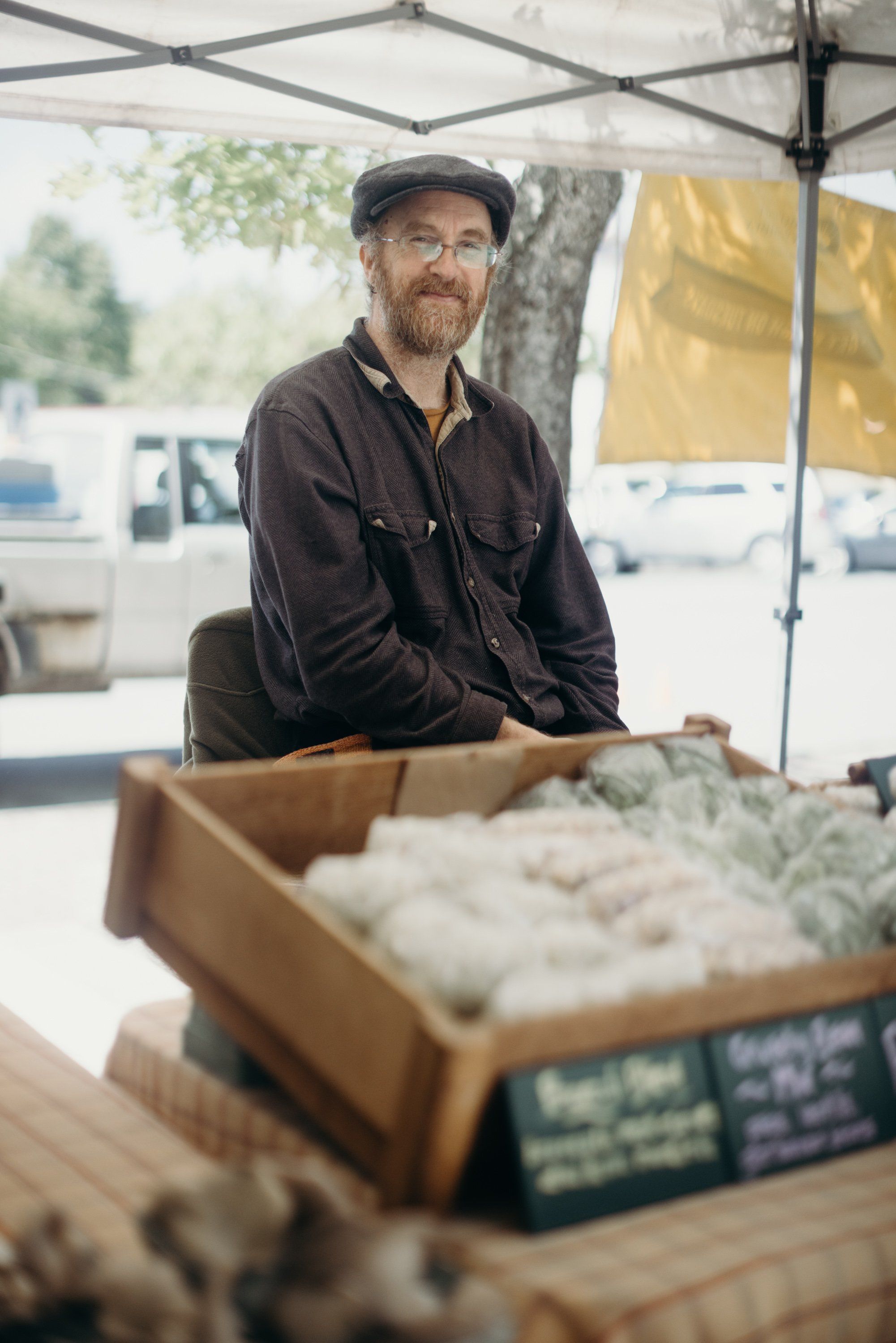 Bearded vendor in cap and dark jacket smiles behind produce boxes at farmers market under tent