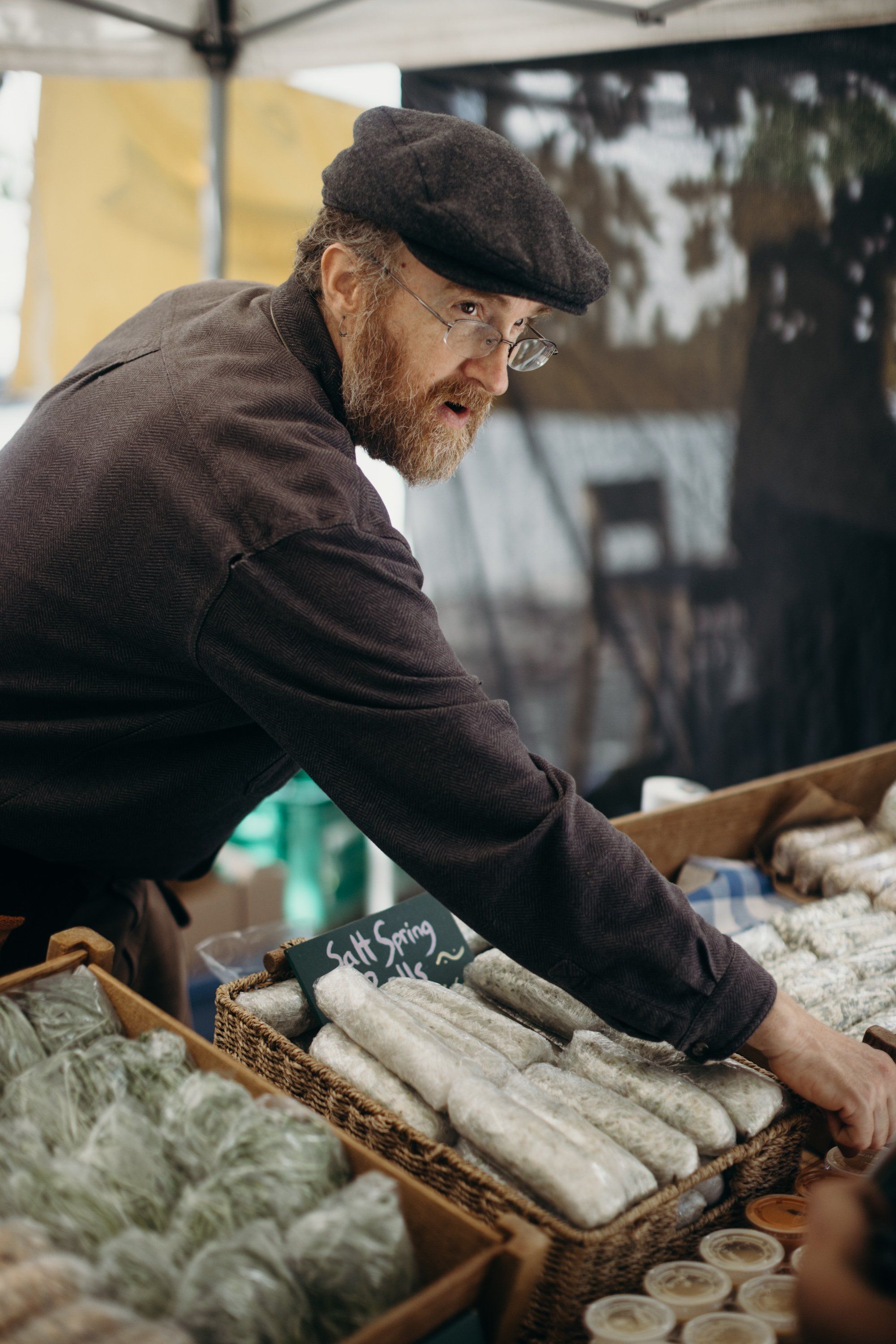 Bearded vendor in brown cap arranges fresh produce at farmers market stand under tent canopy