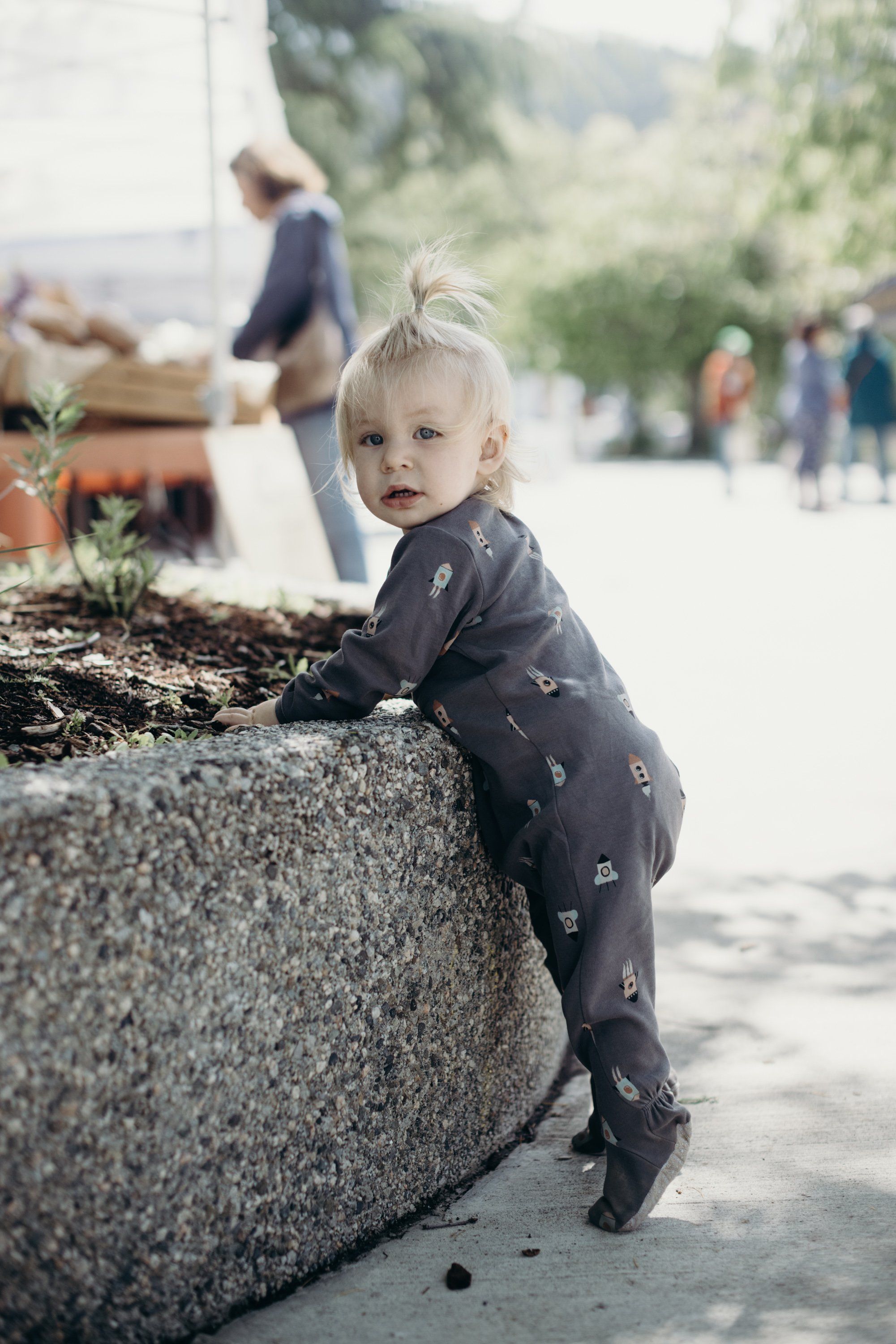 Blonde toddler with windswept hair perches on a weathered stone ledge, wearing a charcoal patterned shirt and gazing thoughtfully toward the camera