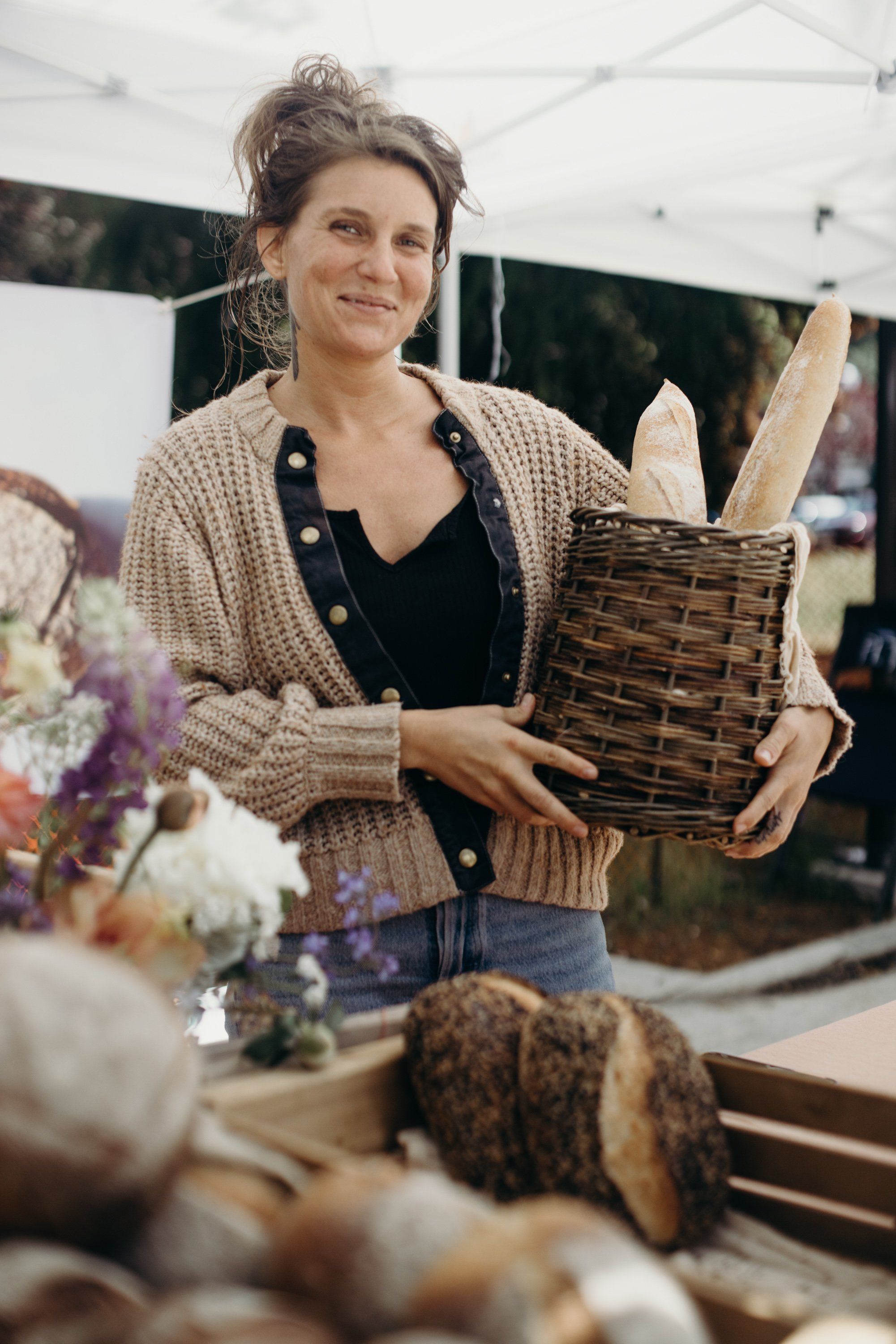 Woman holding woven basket at farmers market with fresh flowers and baked goods displayed nearby