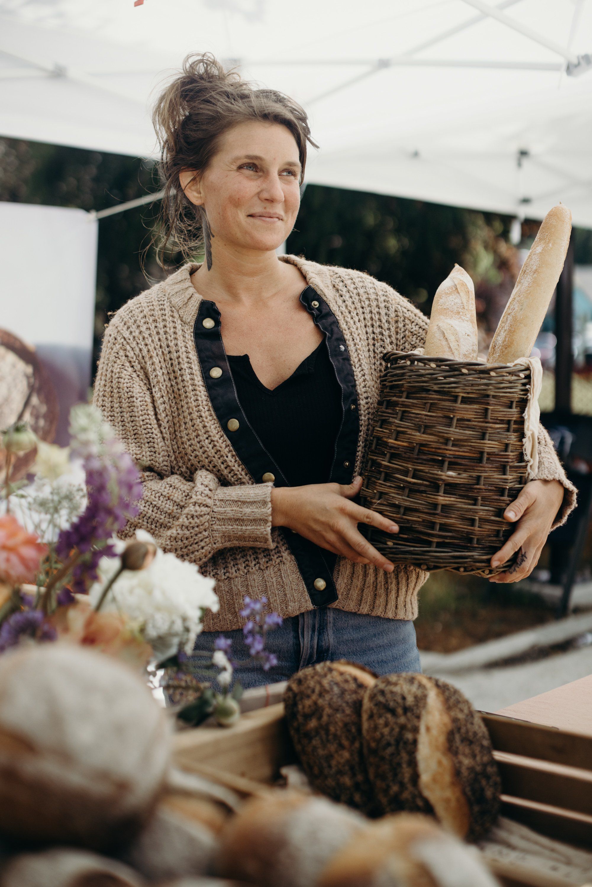 Woman holding woven basket with bread at farmers market, surrounded by fresh flowers and produce in natural light