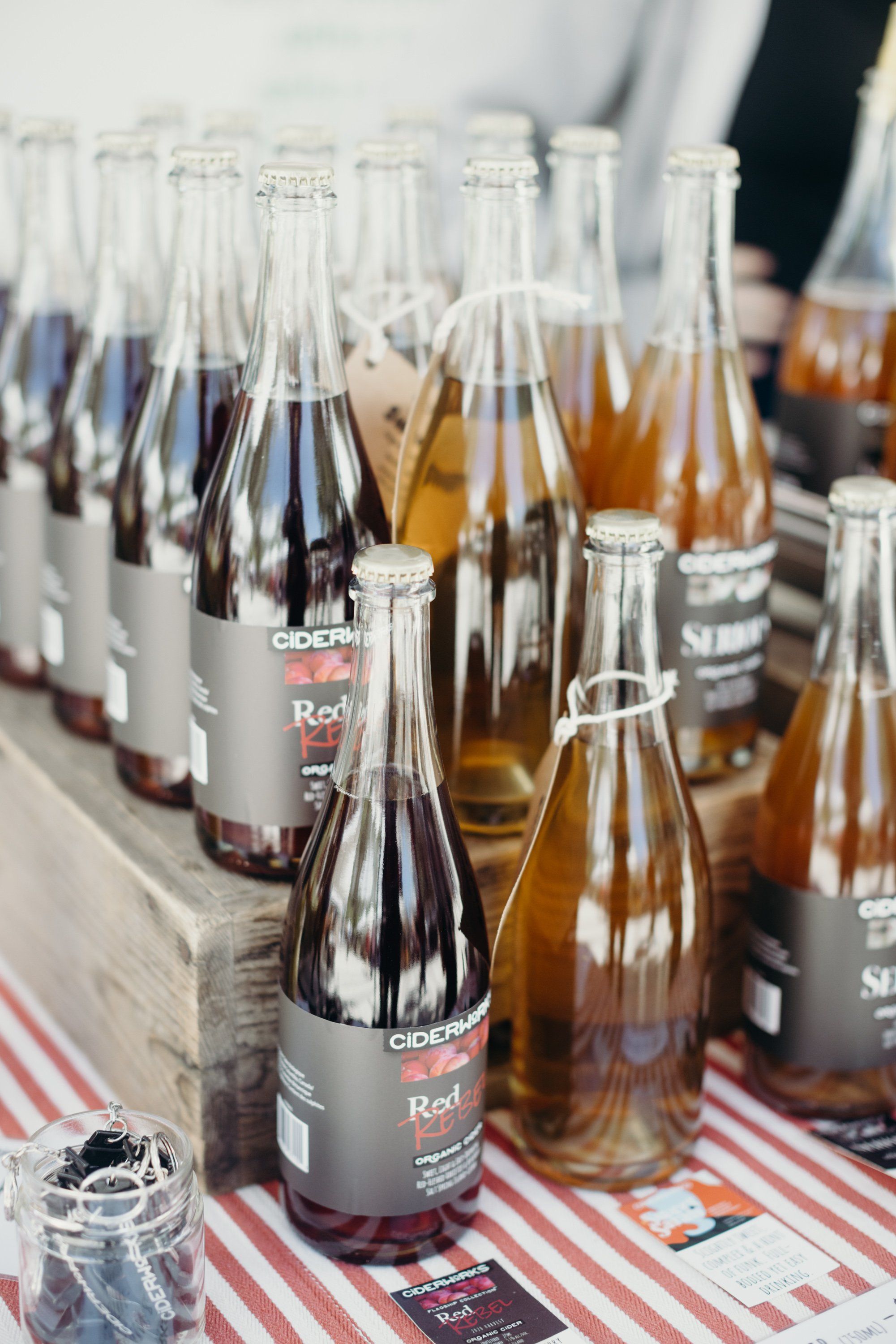 Glass bottles of cider and other beverages lined up on a red and white striped table at an outdoor market