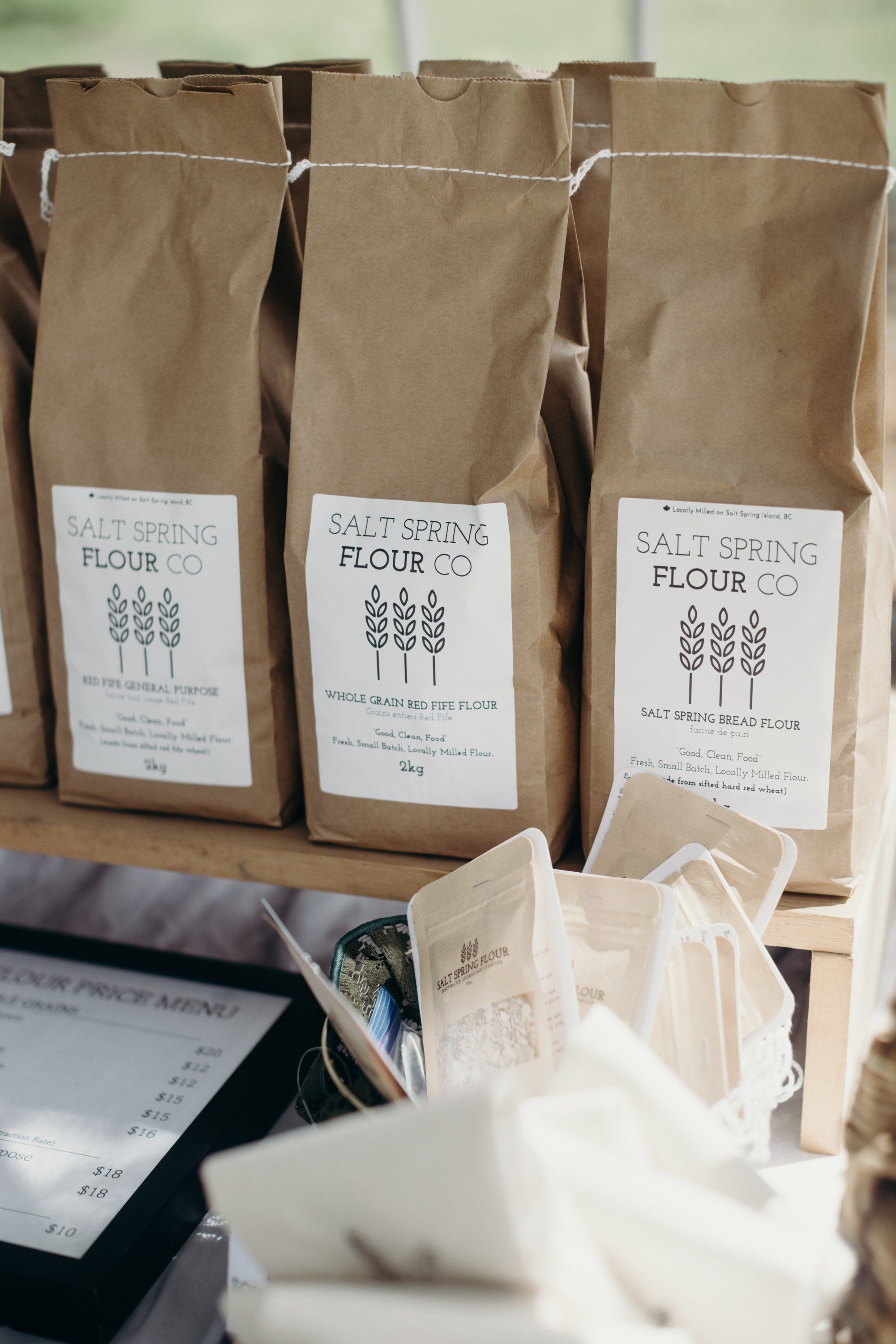 Three kraft paper bags of Salt Spring flour displayed at a farmers market, with white ceramic vessels in soft natural light
