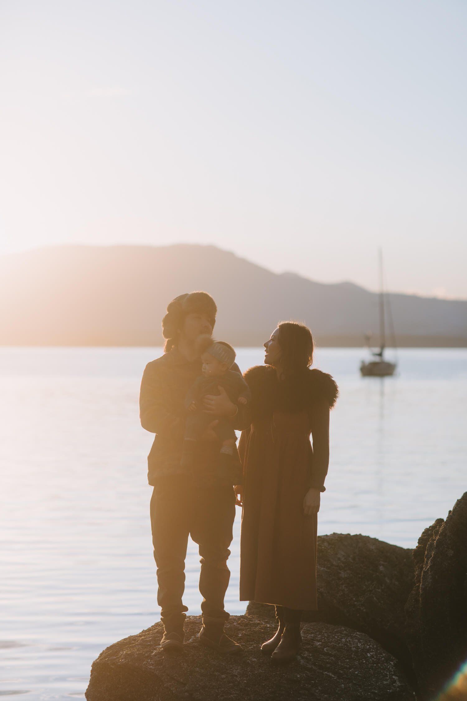 Two children stand on a rocky outcrop at golden hour, gazing toward calm water and distant mountains with a sailboat anchored beyond