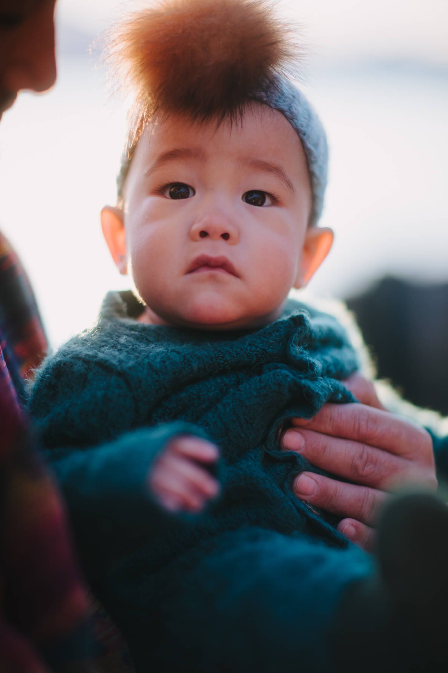 Toddler with tousled reddish hair in blue knit sweater held by parent's hands, soft natural light, gentle and intimate moment