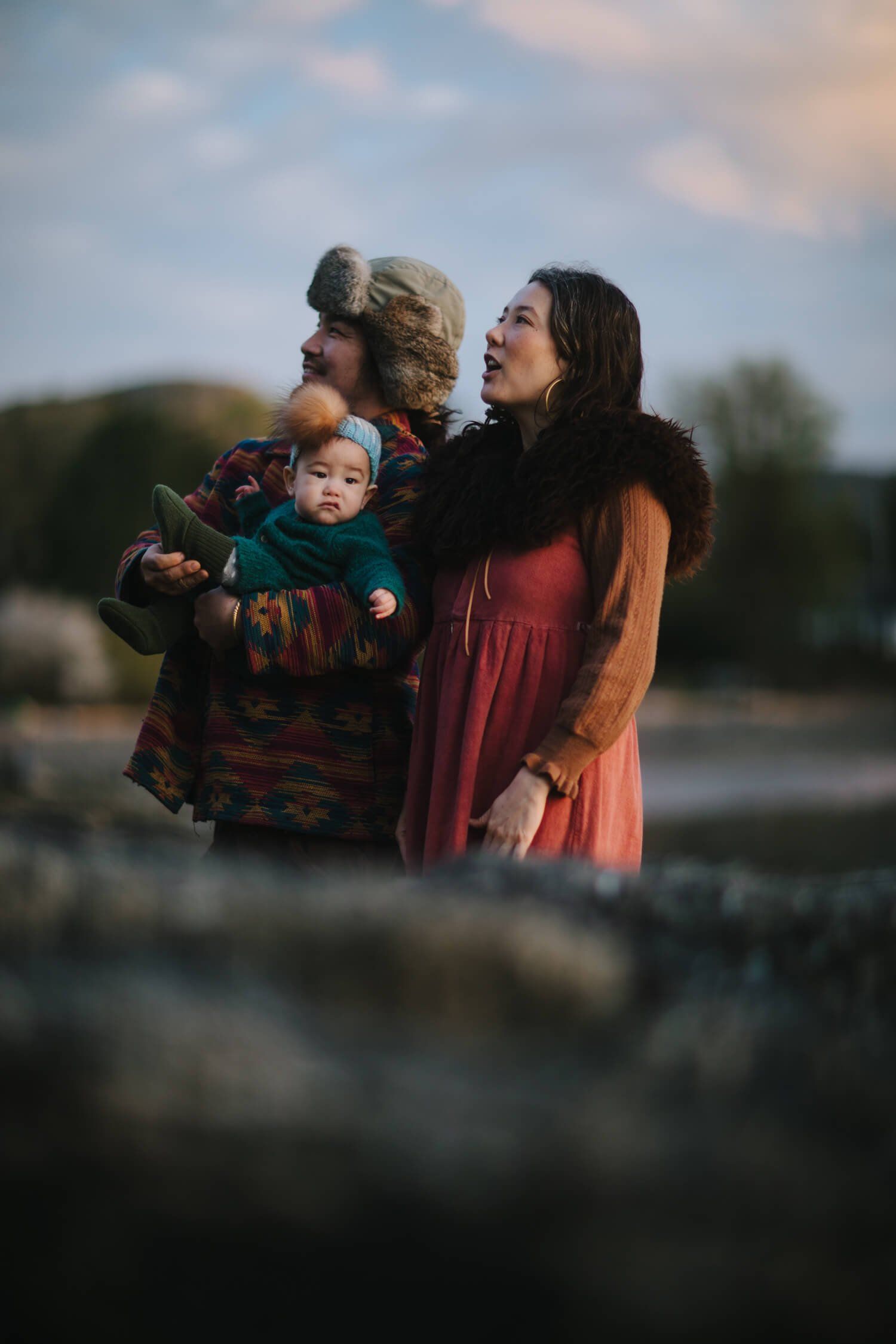 Two parents hold their young child while gazing skyward together near water at golden hour, wrapped in warm autumn clothing