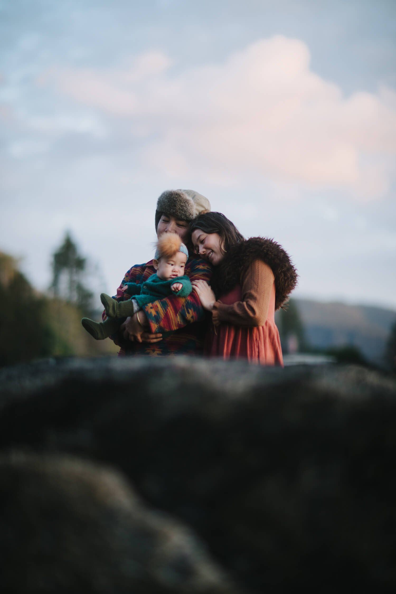 Grandmother holds a toddler while a mother embraces them both outdoors, soft clouds and forested hills glowing behind them