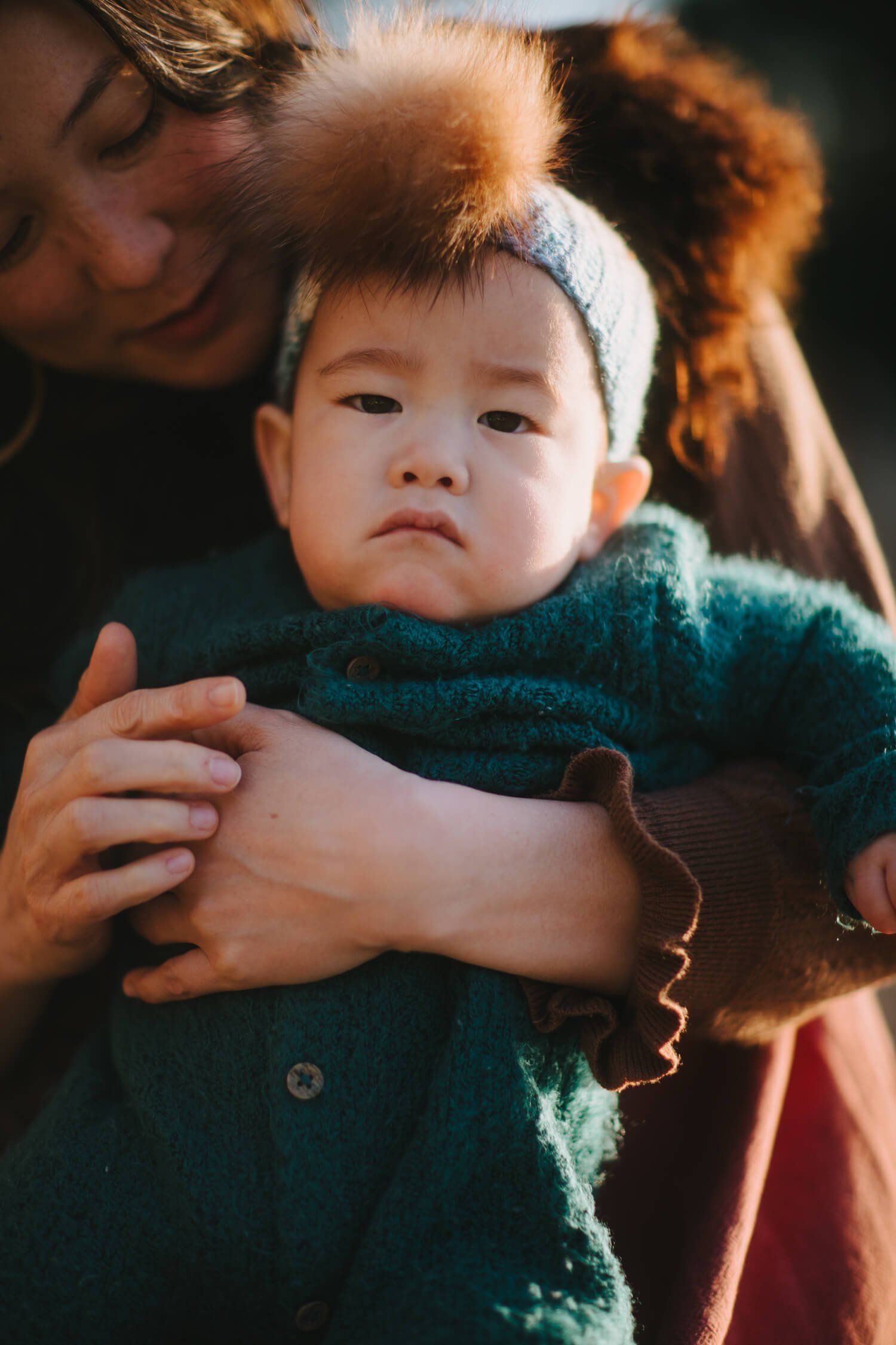 Toddler in forest-green sweater gazes thoughtfully while parent's arms embrace from behind in warm, dappled golden light