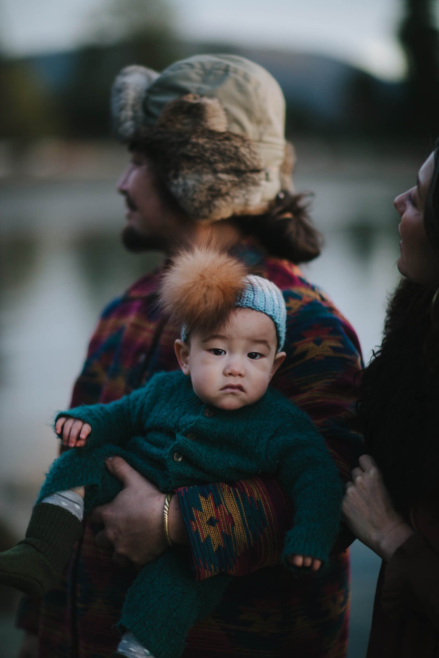 Toddler in teal knit sweater and blue hat held by parent in plaid shirt beside water, soft natural light