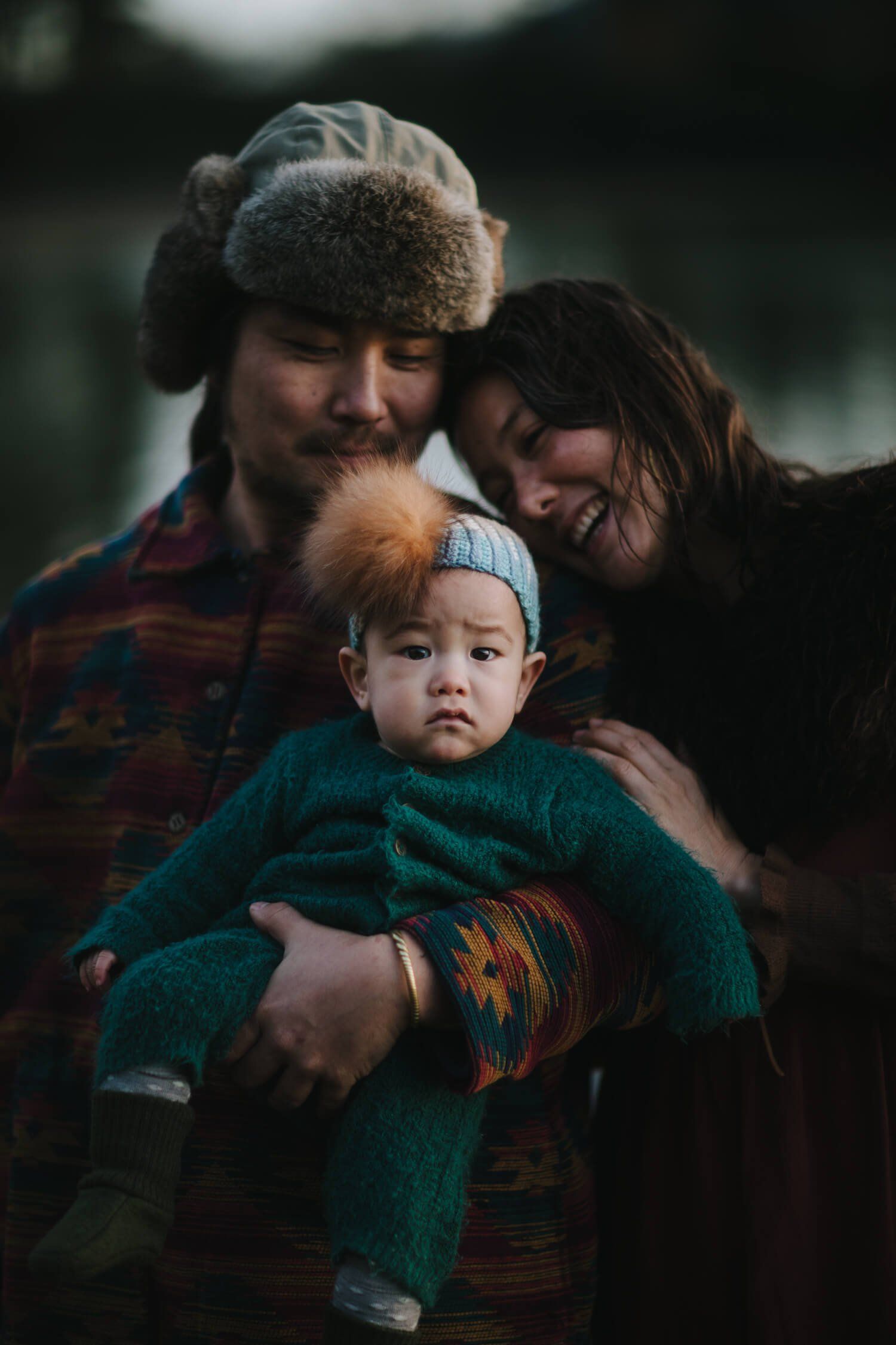 Young family of three embraced outdoors in soft golden light, parents smiling down at bundled infant in teal sweater and knit hat