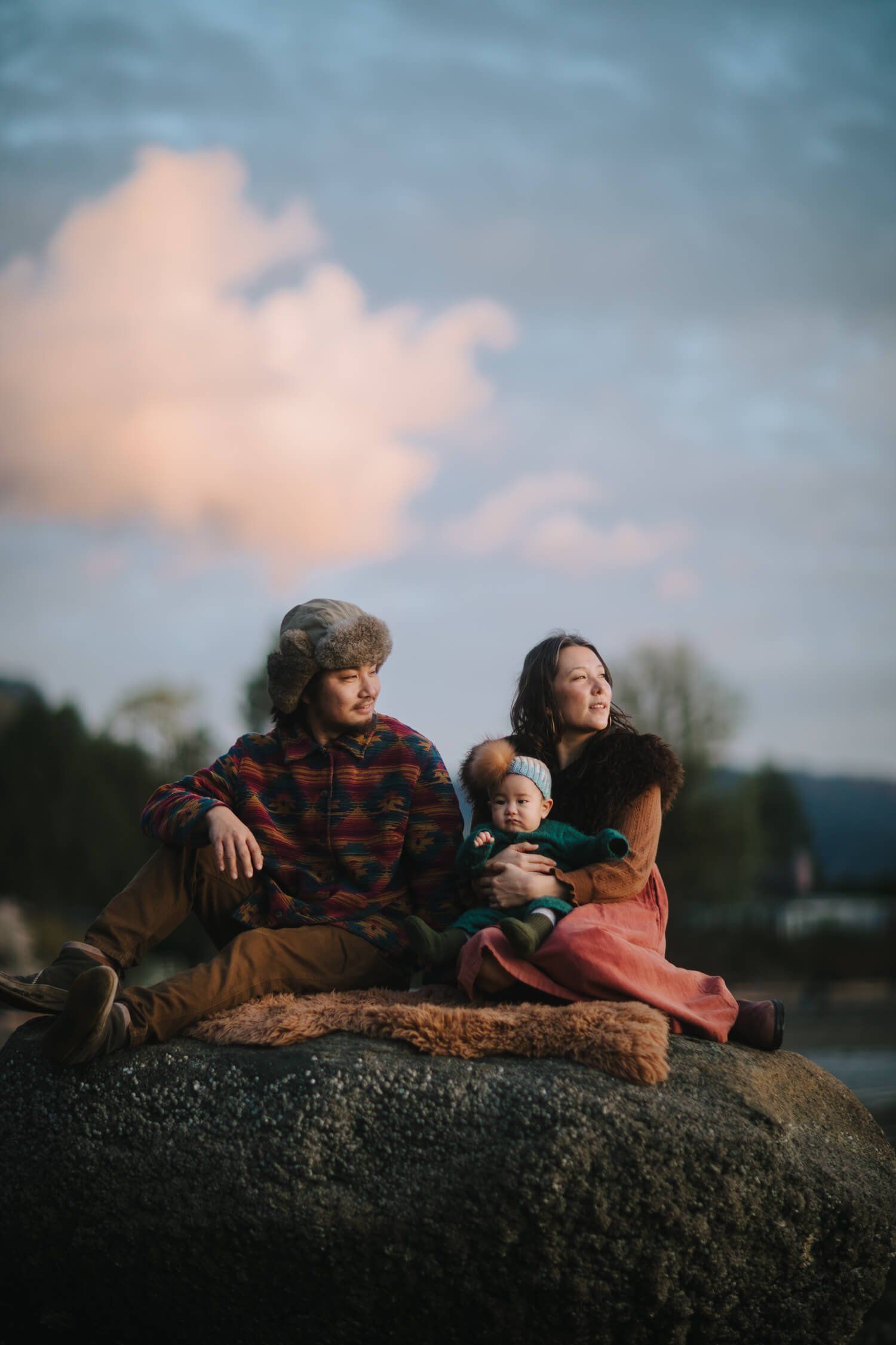 Family of three sitting on a boulder at golden hour, overlooking water with forested shores and soft pink clouds above
