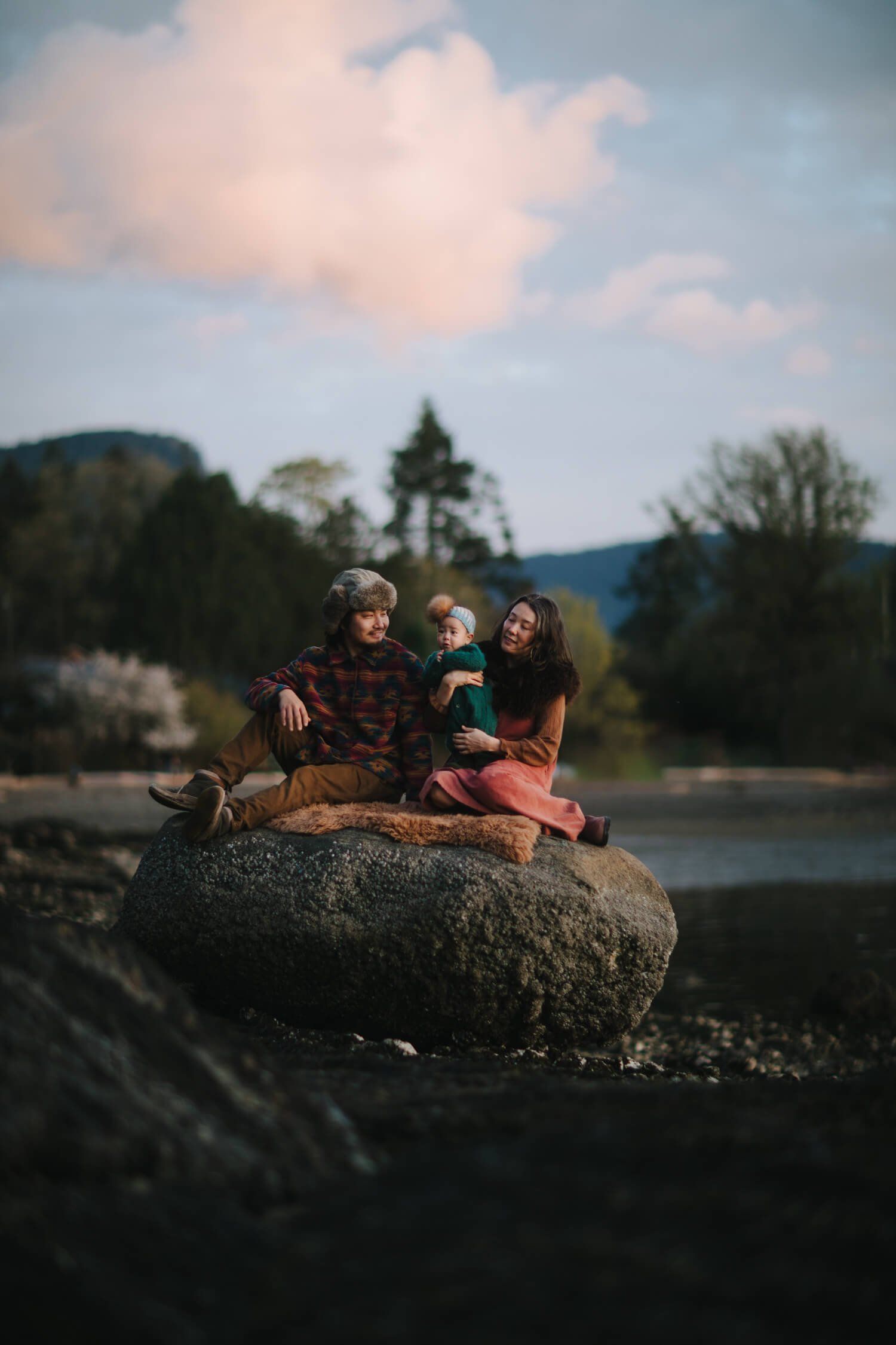 Three people sit on a large boulder by a river with forested mountains and soft evening light in the background