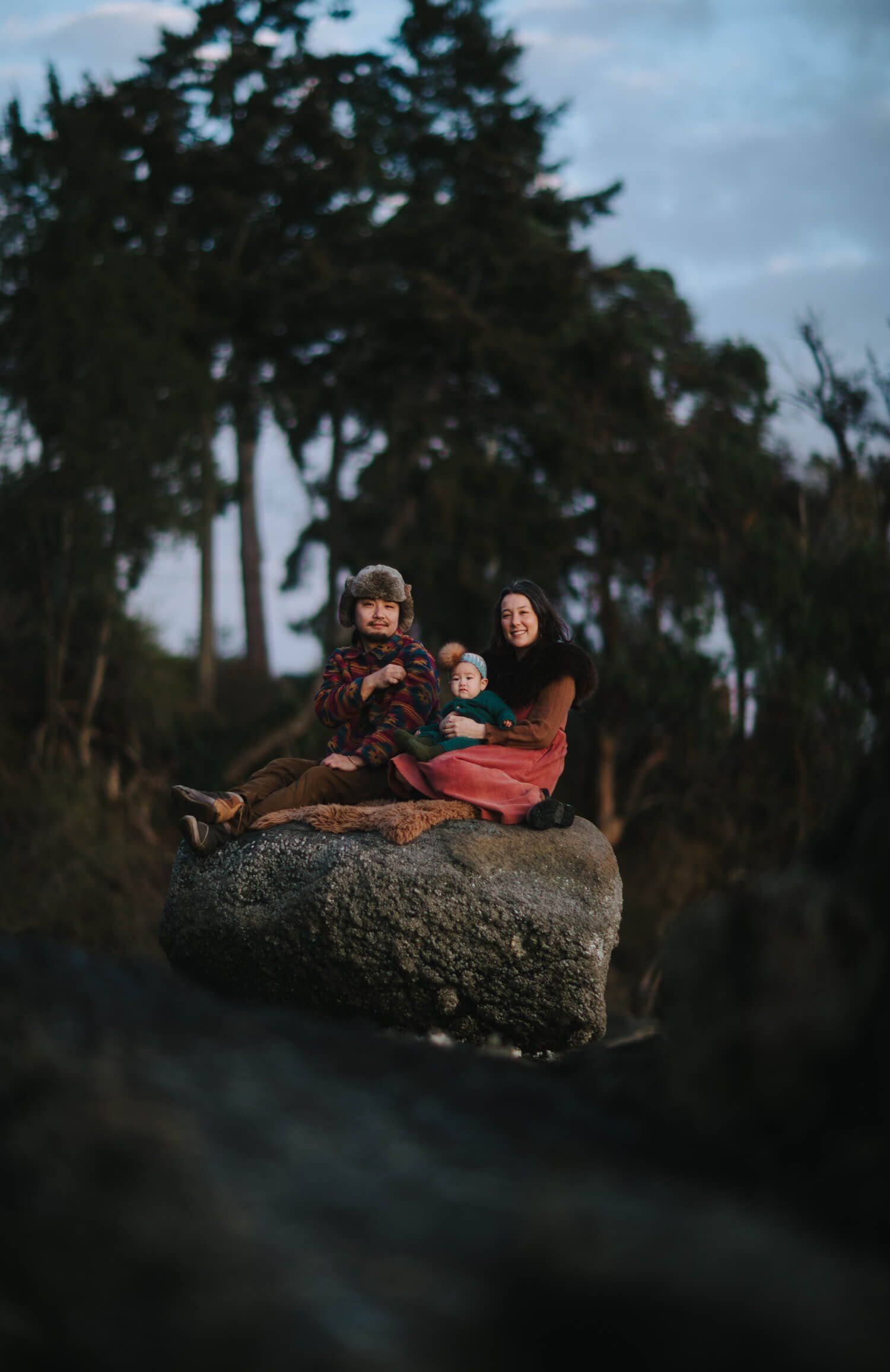 Pregnant woman and child sit on a moss-covered boulder surrounded by tall evergreen trees in soft, overcast light