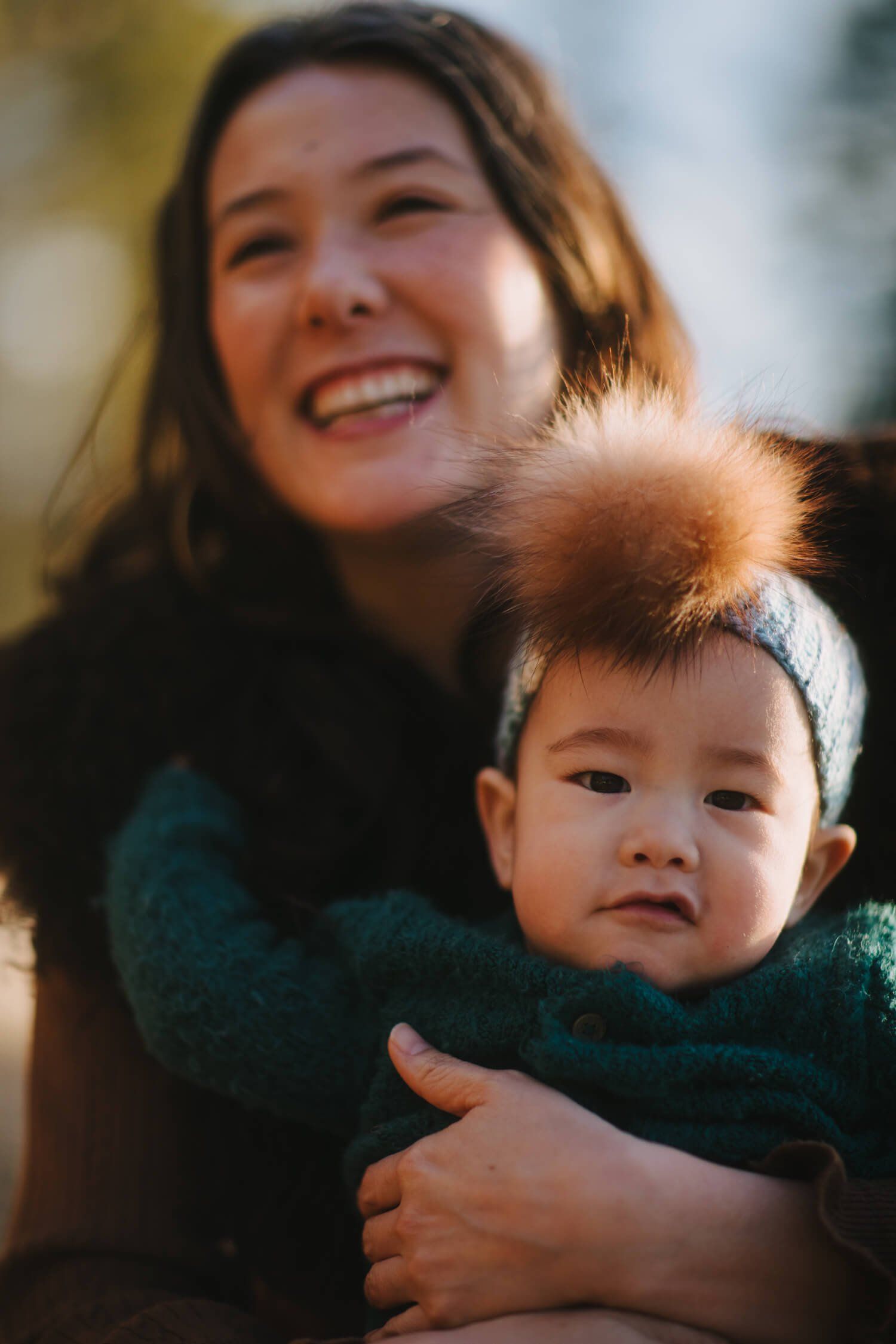 Woman in dark sweater smiling warmly while holding a toddler with fluffy hair, bathed in golden sunlight