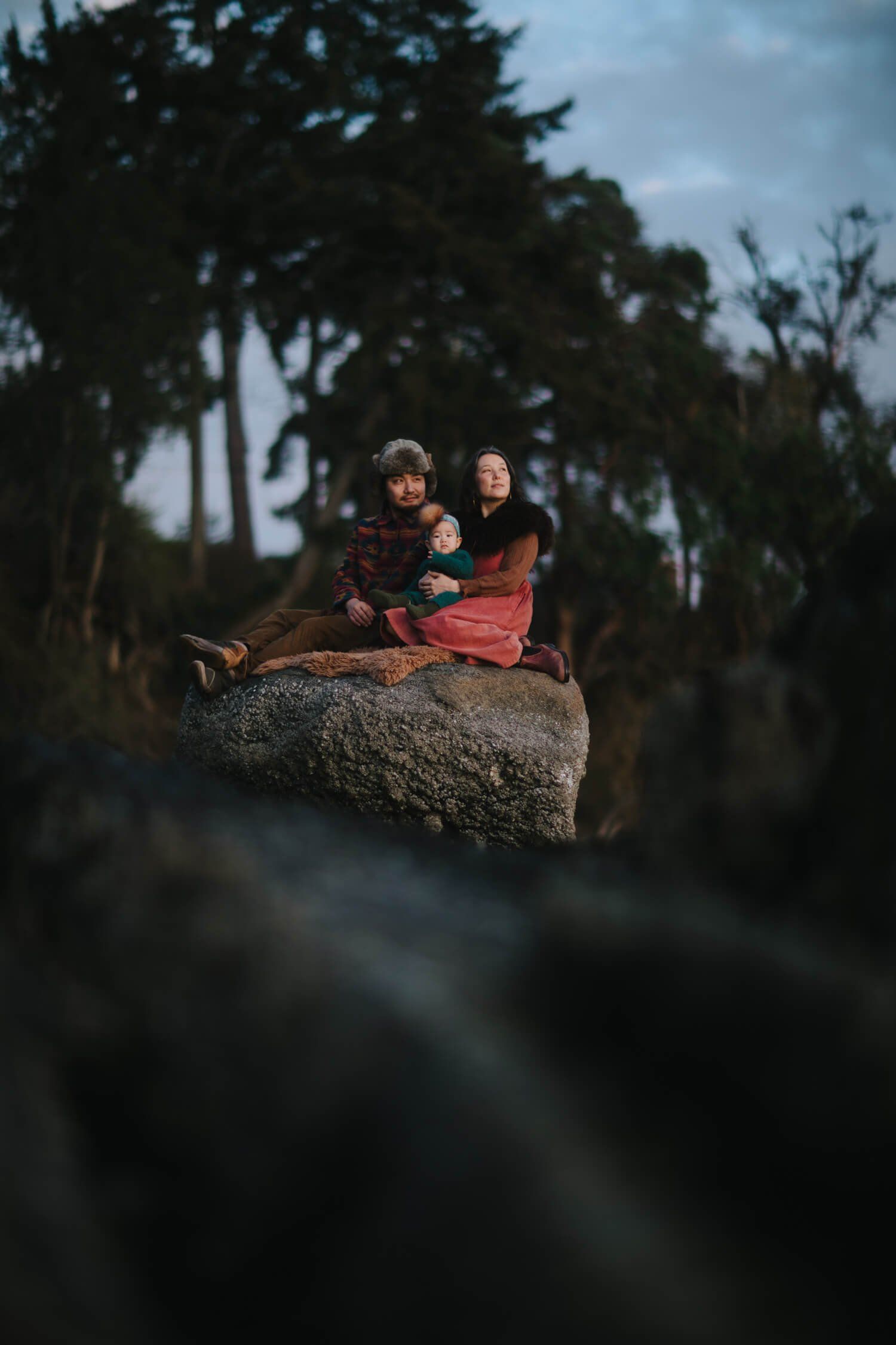 Expectant parents sit together on a moss-covered boulder beneath towering evergreens at dusk, glowing warmly in soft amber light