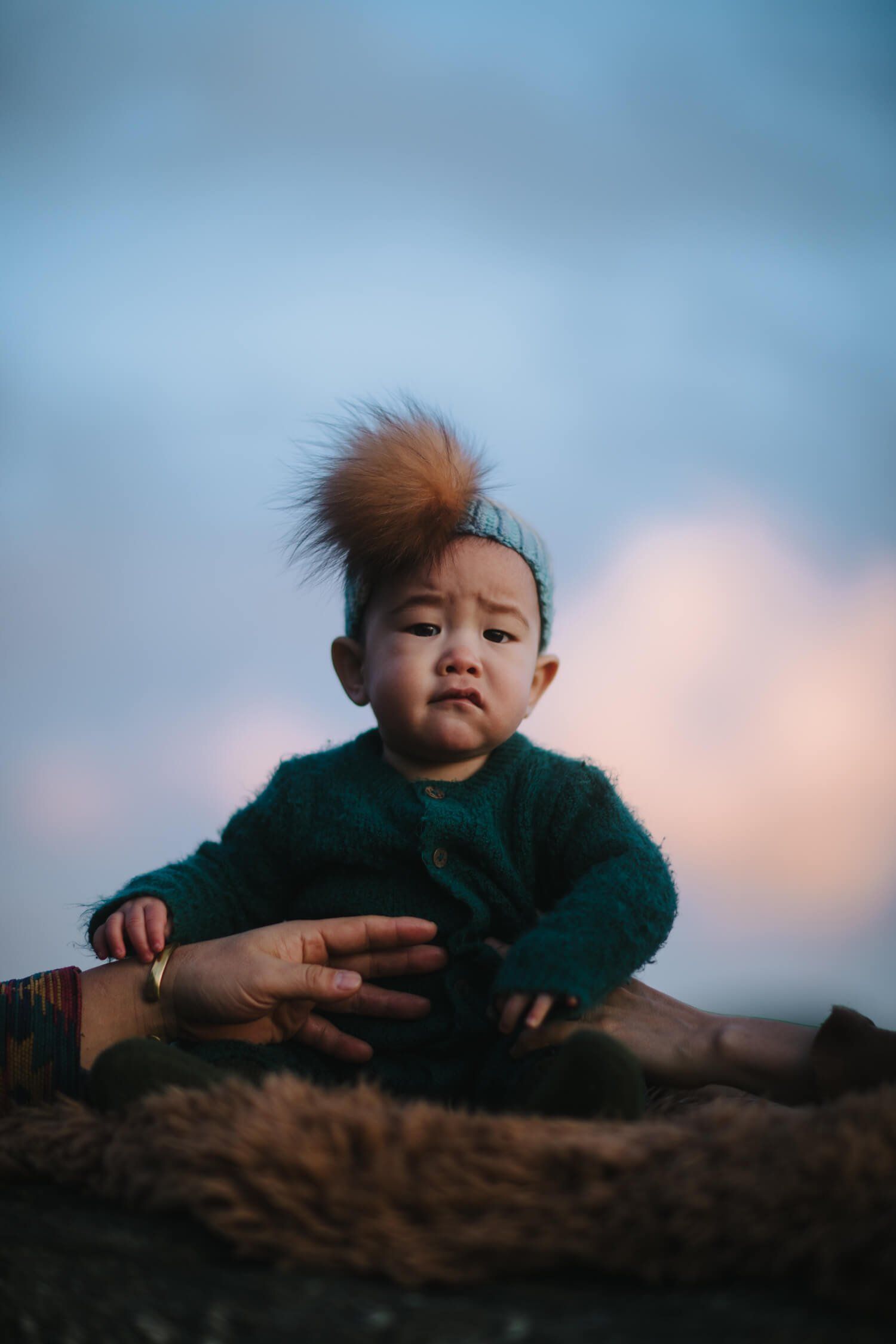 Toddler in forest-green sweater sits on a log against a dusky blue sky, gazing thoughtfully to the side with tousled hair catching warm light