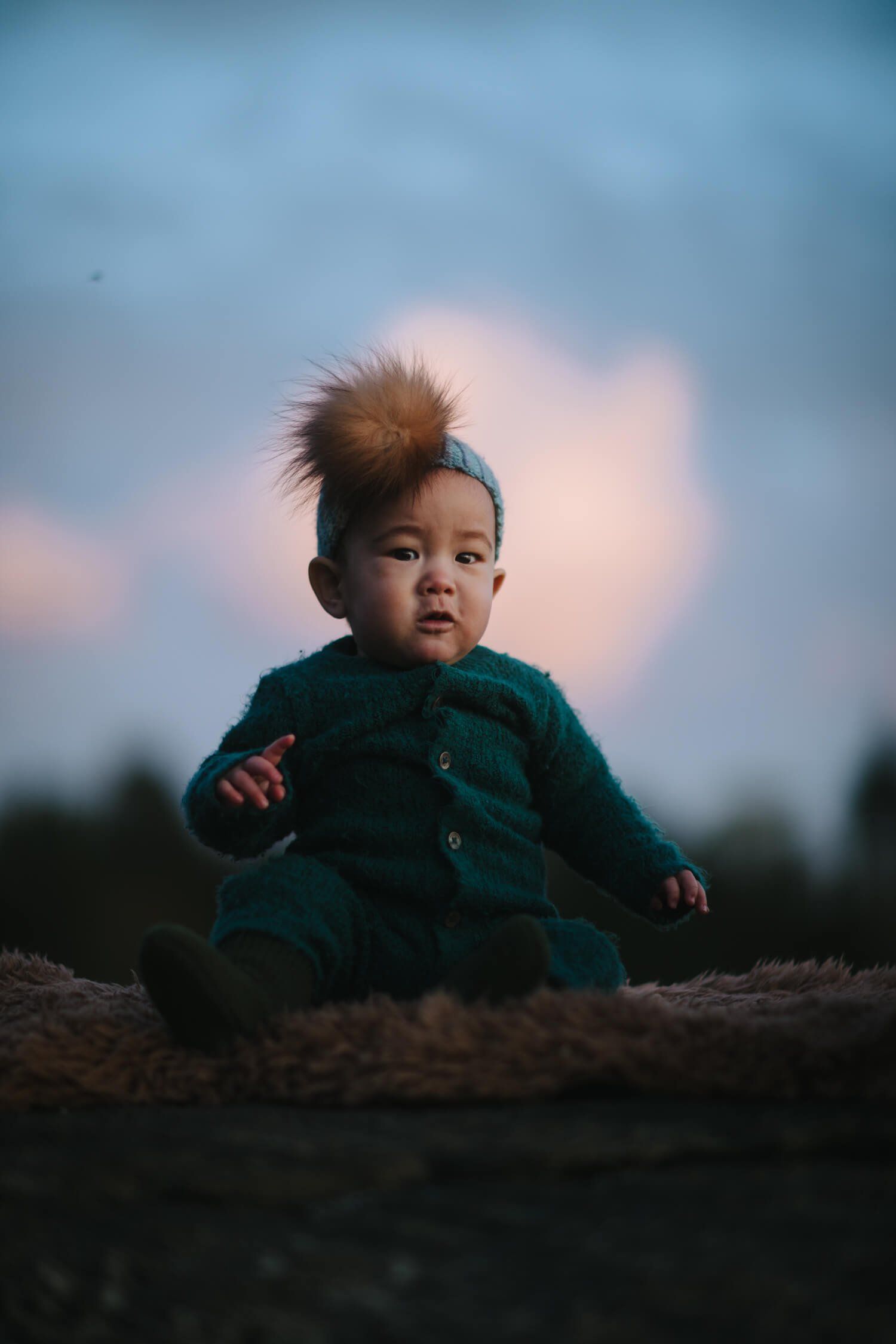 Toddler in teal jacket sits on dark earth at dusk, backlit by soft pink and blue sky, gazing thoughtfully to the side