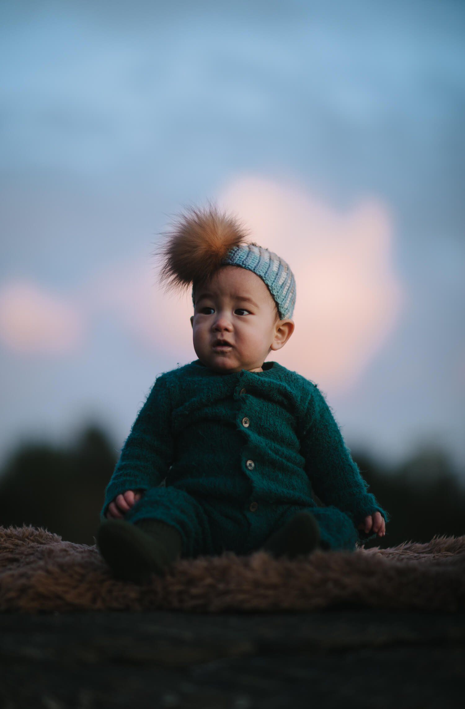 Toddler in teal cardigan and knit headband sits on driftwood at dusk, gazing upward with soft golden light on their face