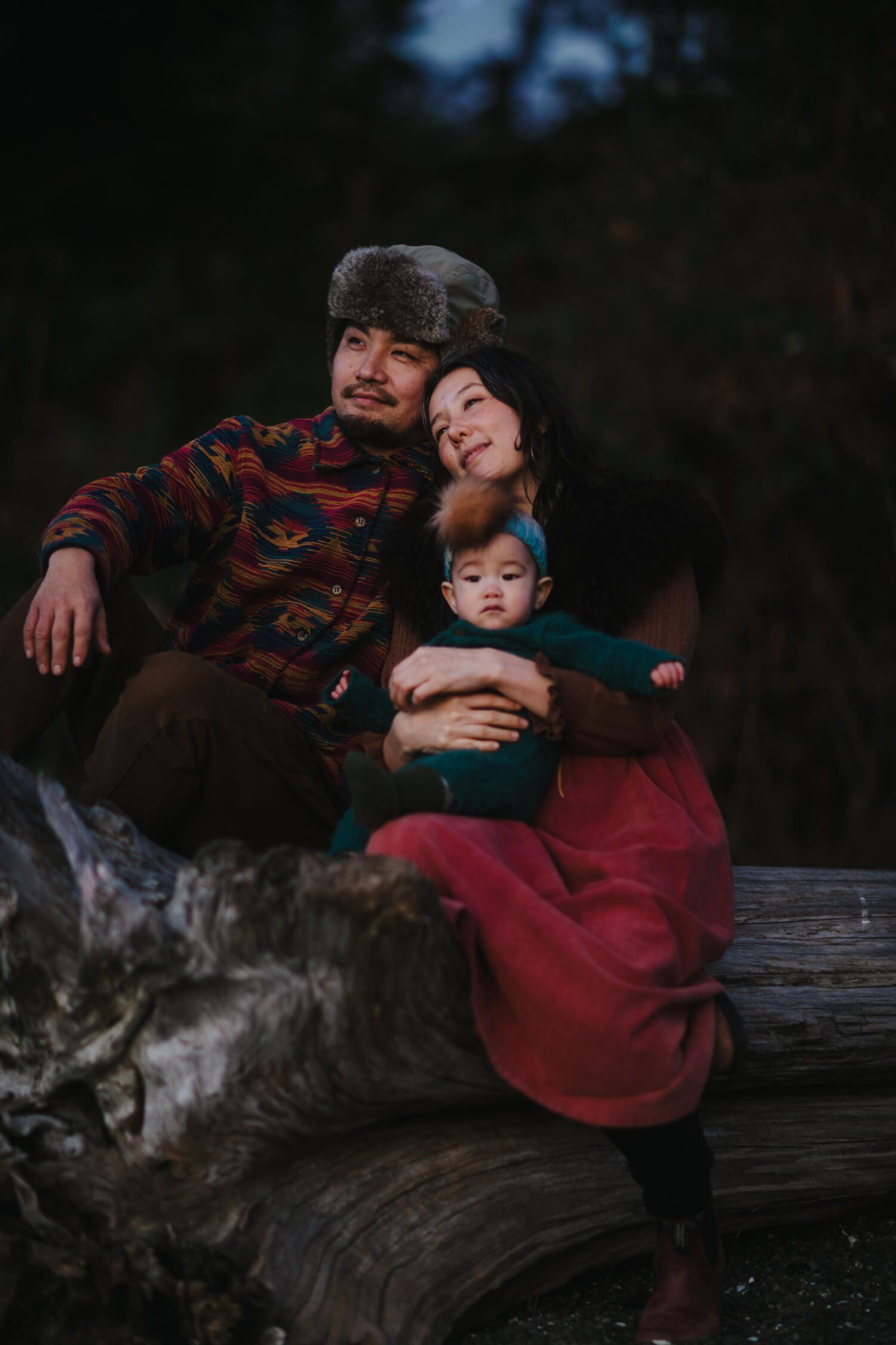 Family of three nestled on a weathered log in moody forest light, wearing warm earth tones and burgundy