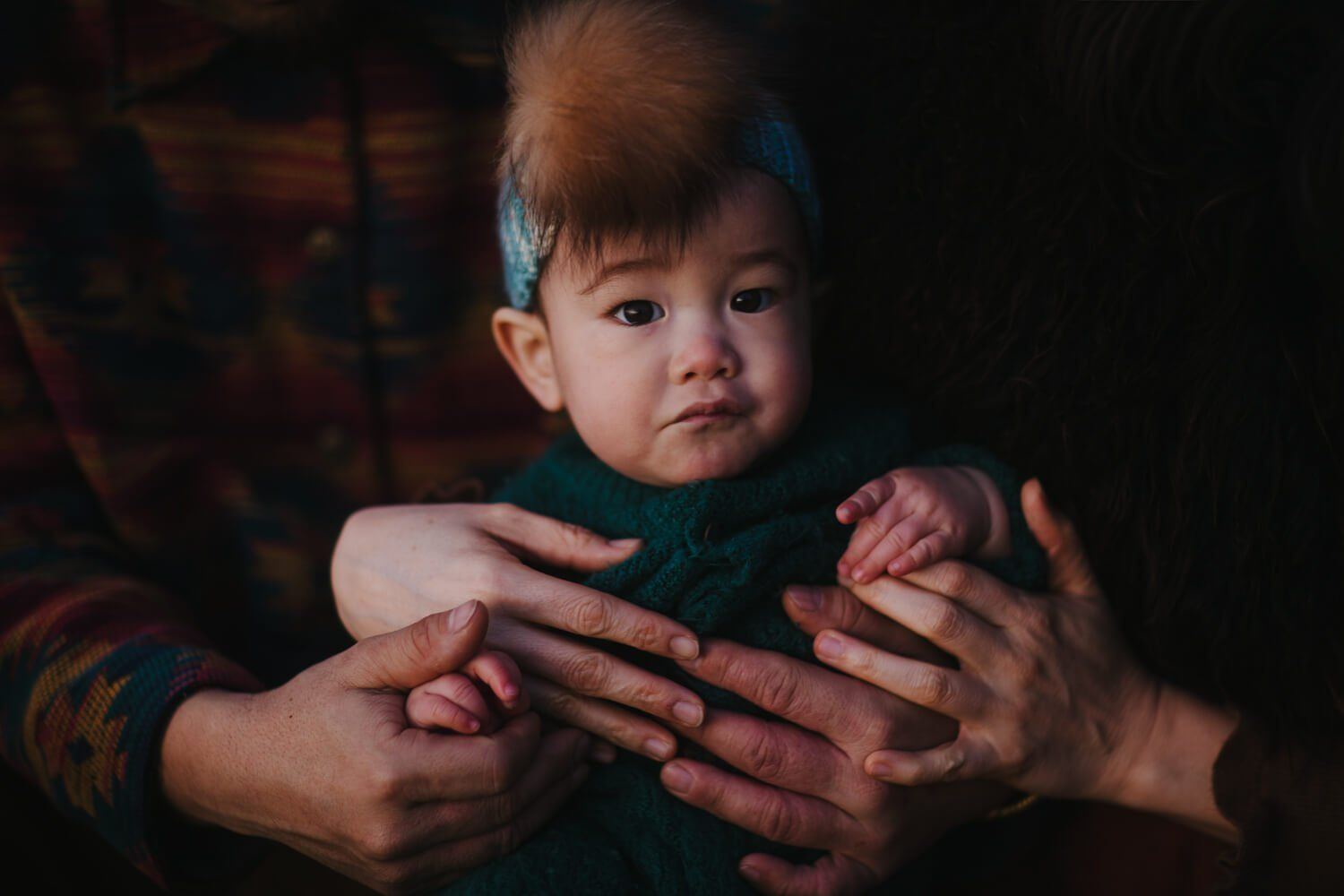 Newborn cradled in parent's hands against dark background, warm light illuminating baby's curious expression and tender skin-to-skin contact