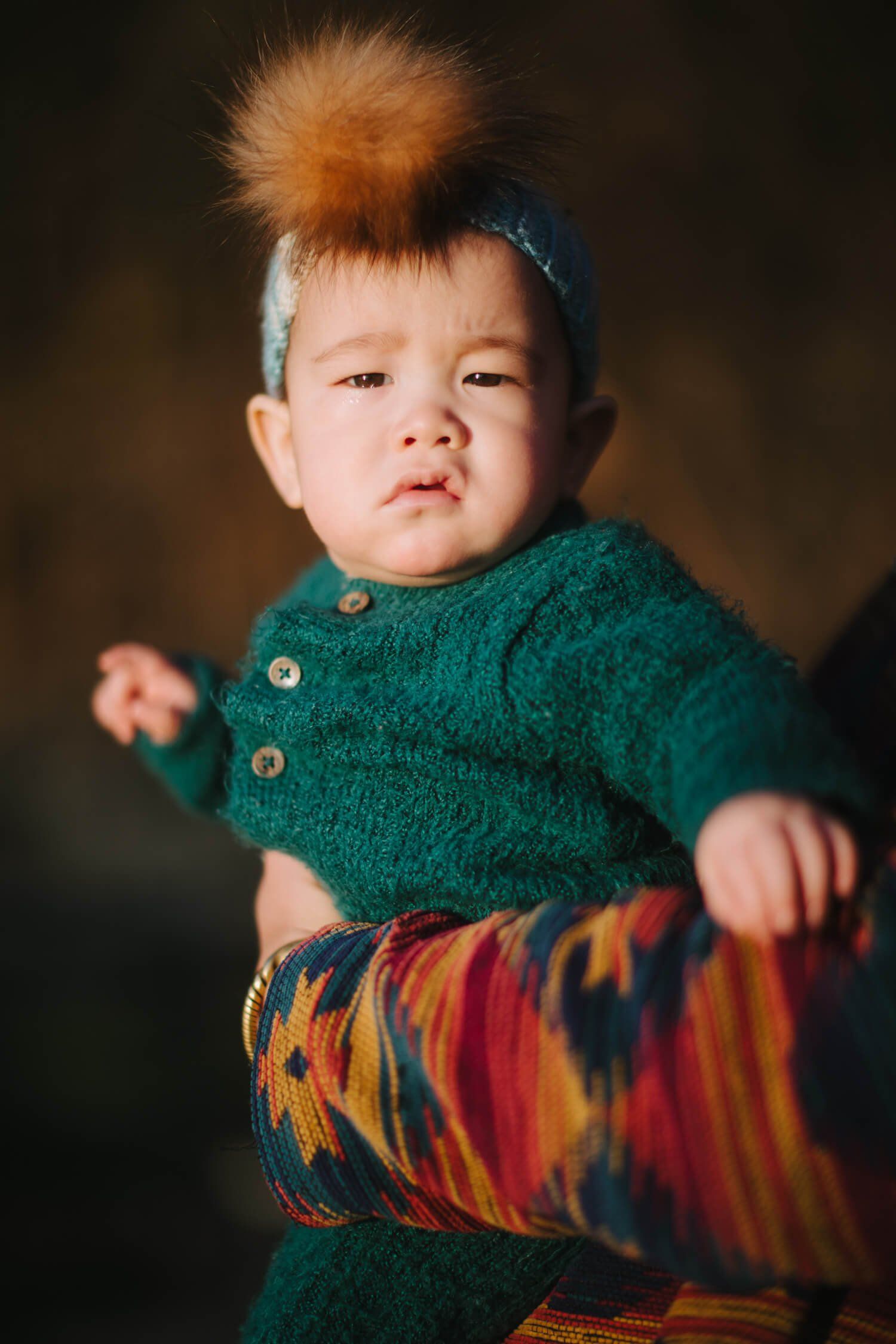 Toddler with spiky auburn hair wearing a teal cardigan and striped blanket against a soft brown backdrop