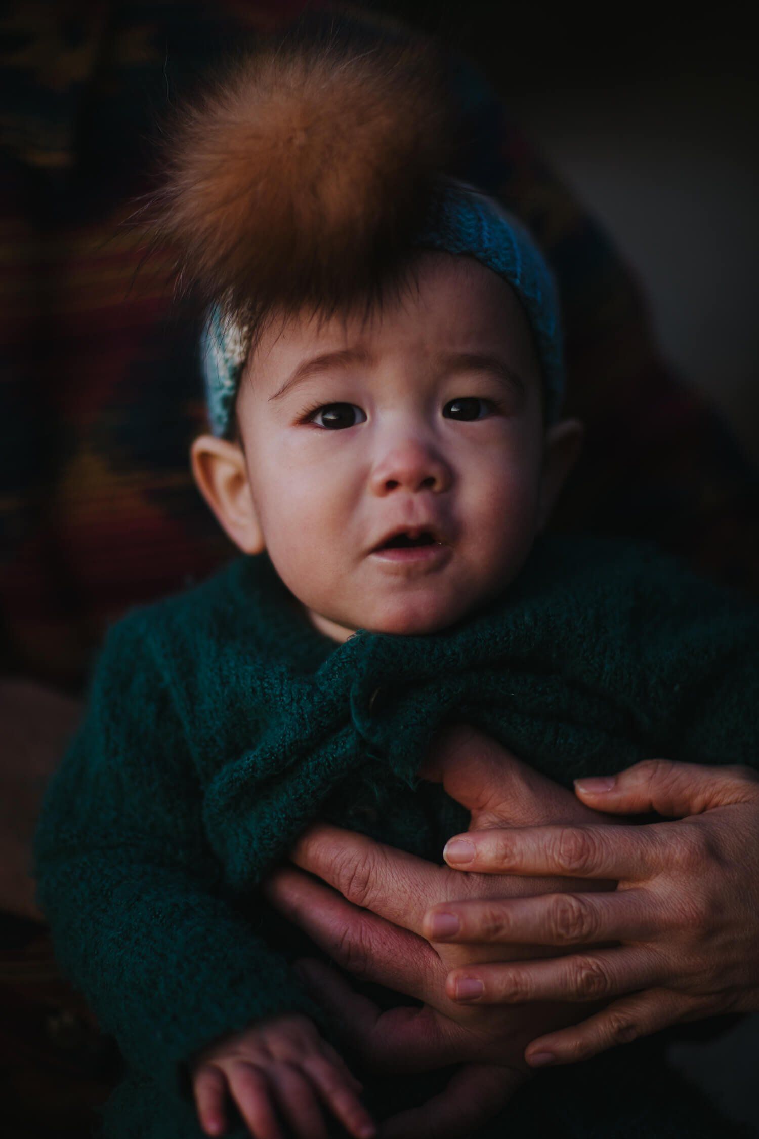 Toddler in teal clothing cradled in parent's hands against dark background, soft warm light illuminating round face and gentle expression
