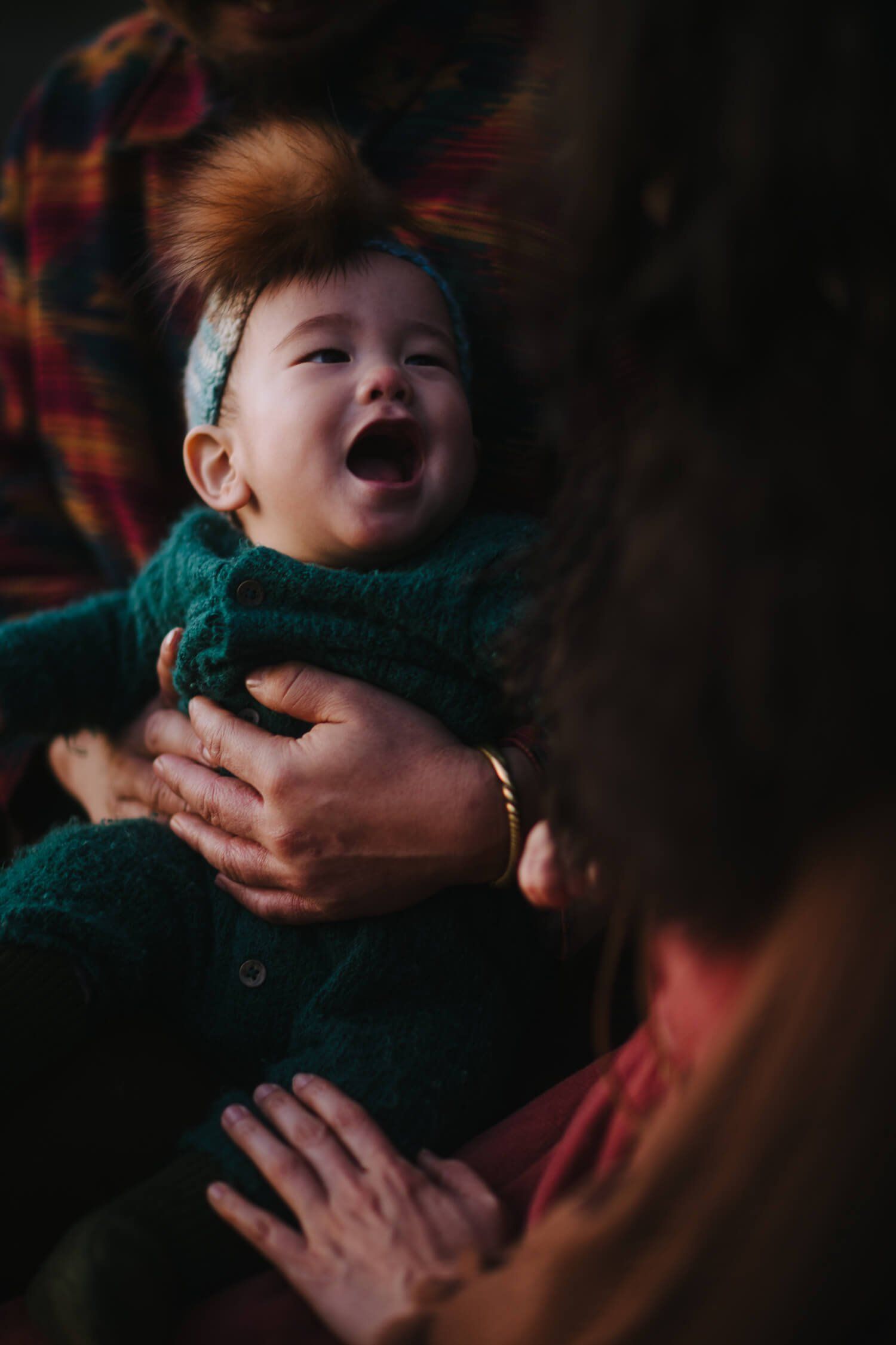 Infant in teal sweater laughs with mouth open while held by parent in warm, intimate indoor lighting