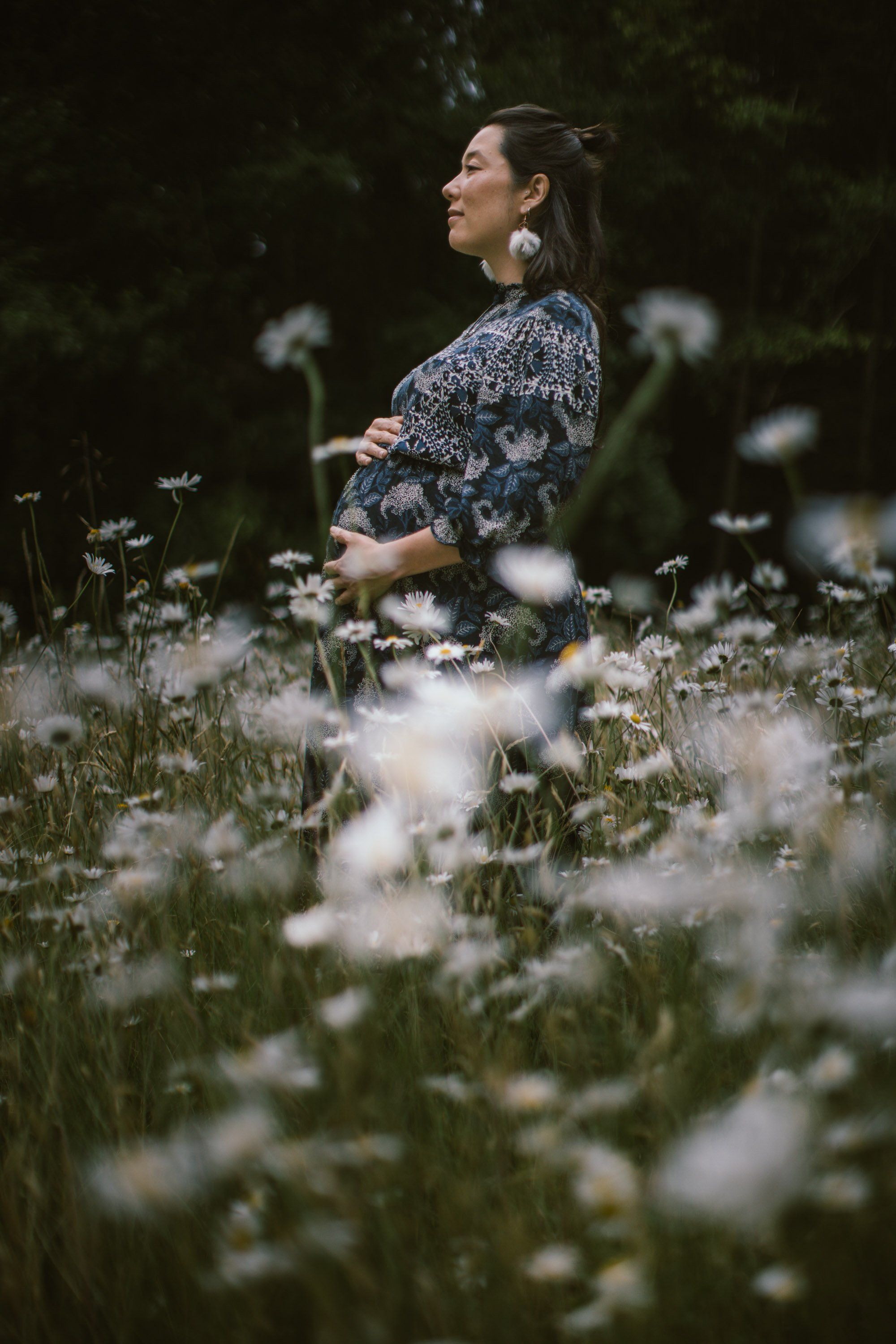 Pregnant woman in patterned blue shirt gazes thoughtfully among blooming white flowers in soft, moody natural light