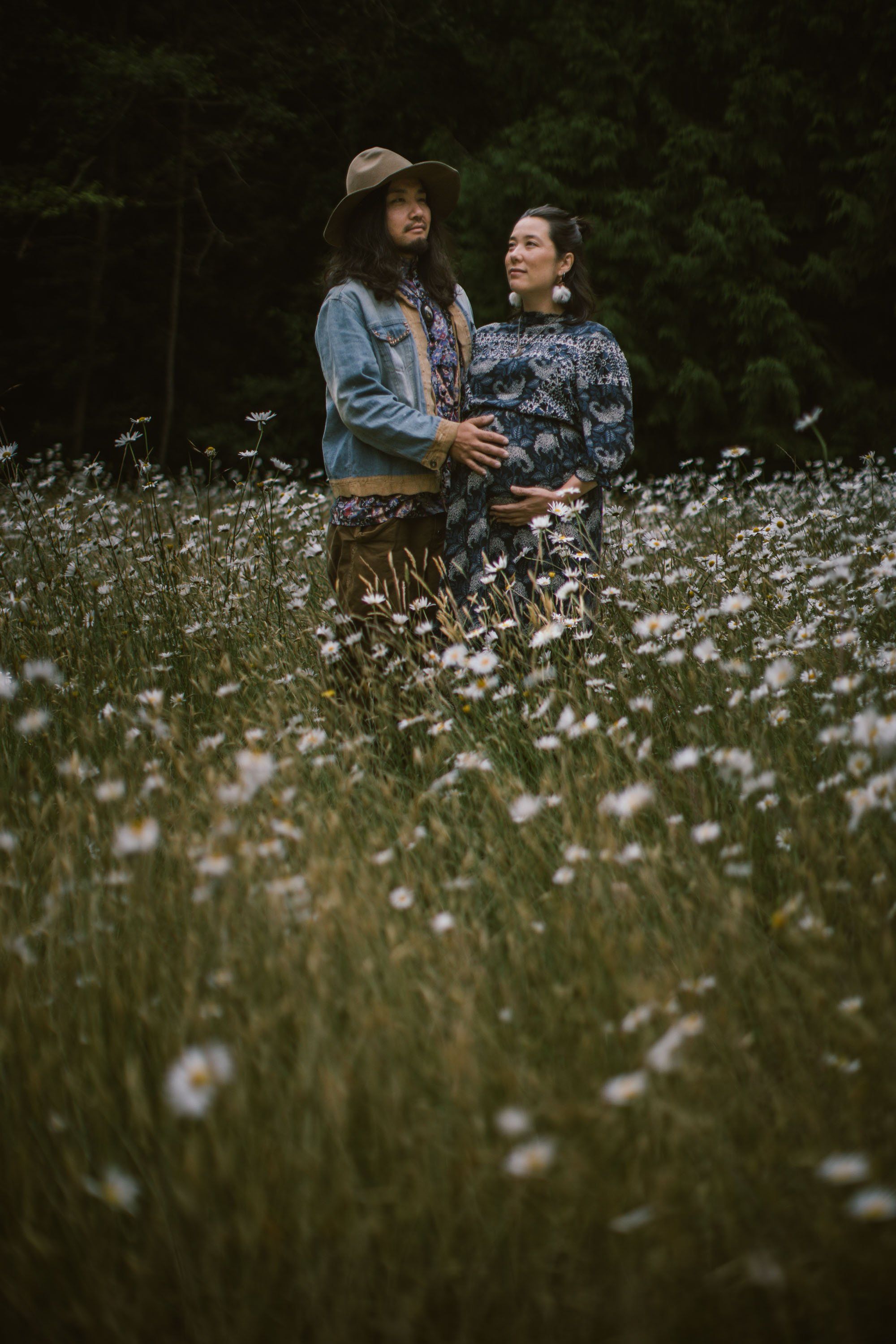 Expecting couple standing amid wildflowers at dusk, soft natural light catching white blossoms in moody forest setting