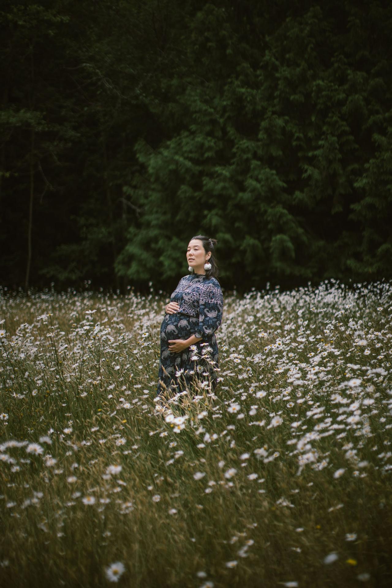 Pregnant woman stands among white wildflowers with evergreen forest backdrop on Salt Spring Island