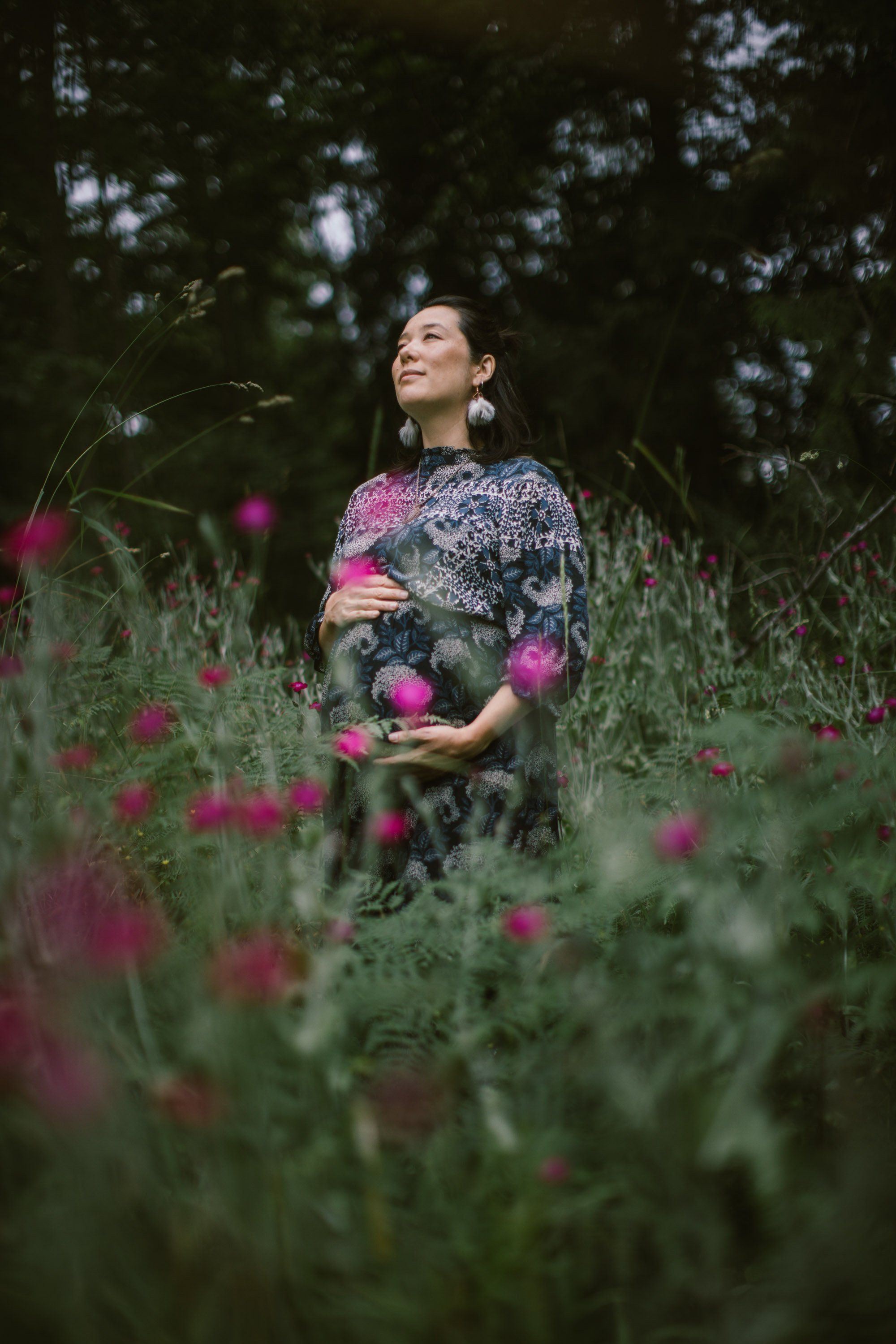 Pregnant woman in a patterned sweater gazes upward amid pink thistle flowers in a sun-dappled forest garden
