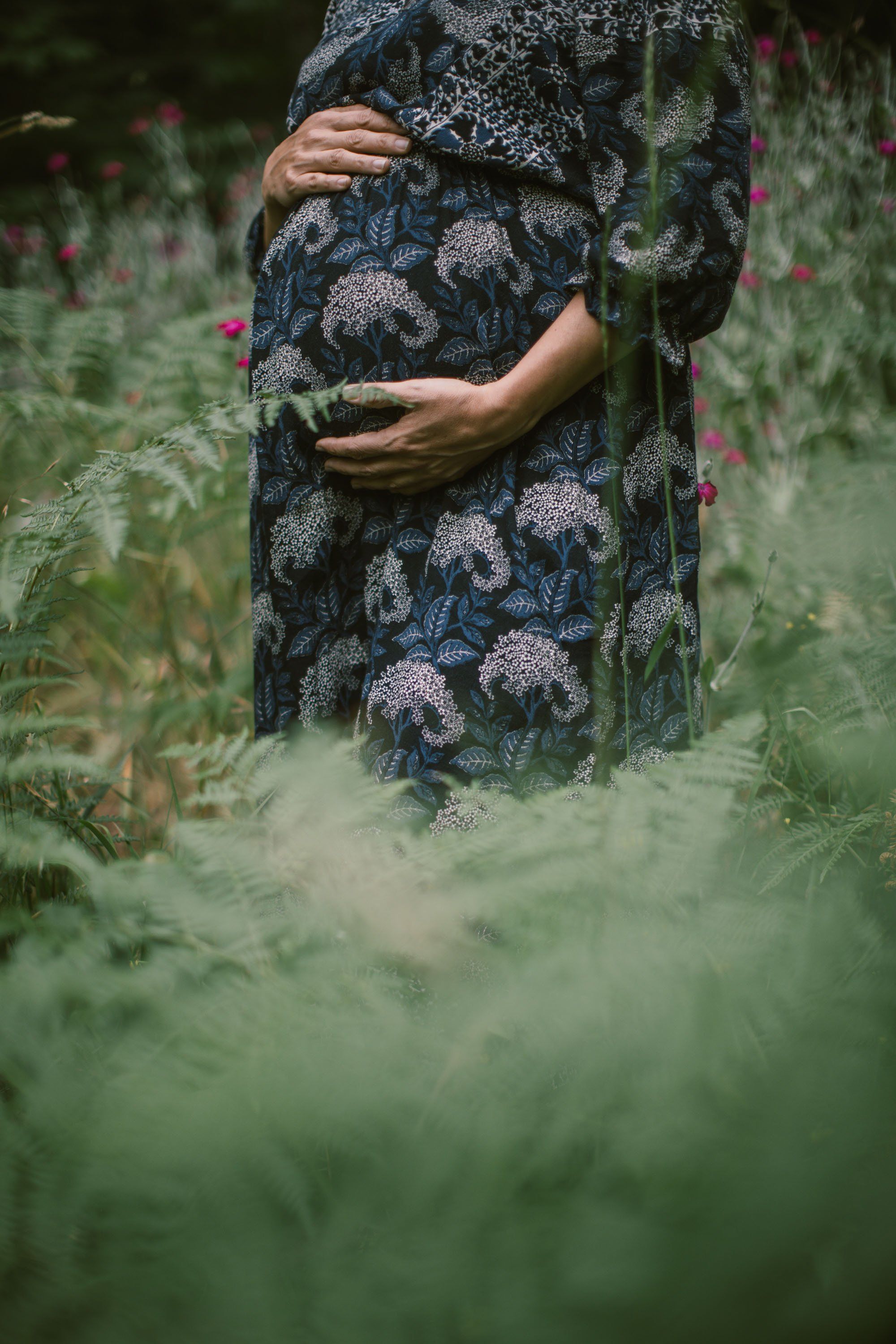 Pregnant person cradling belly in patterned dress among tall green garden plants and purple flowers