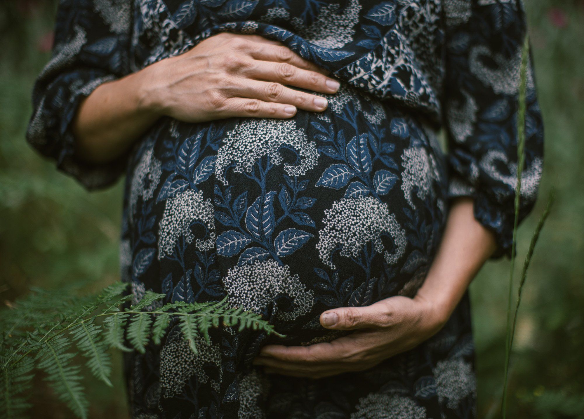Pregnant woman in patterned dress cradling belly with both hands among green ferns, soft natural light