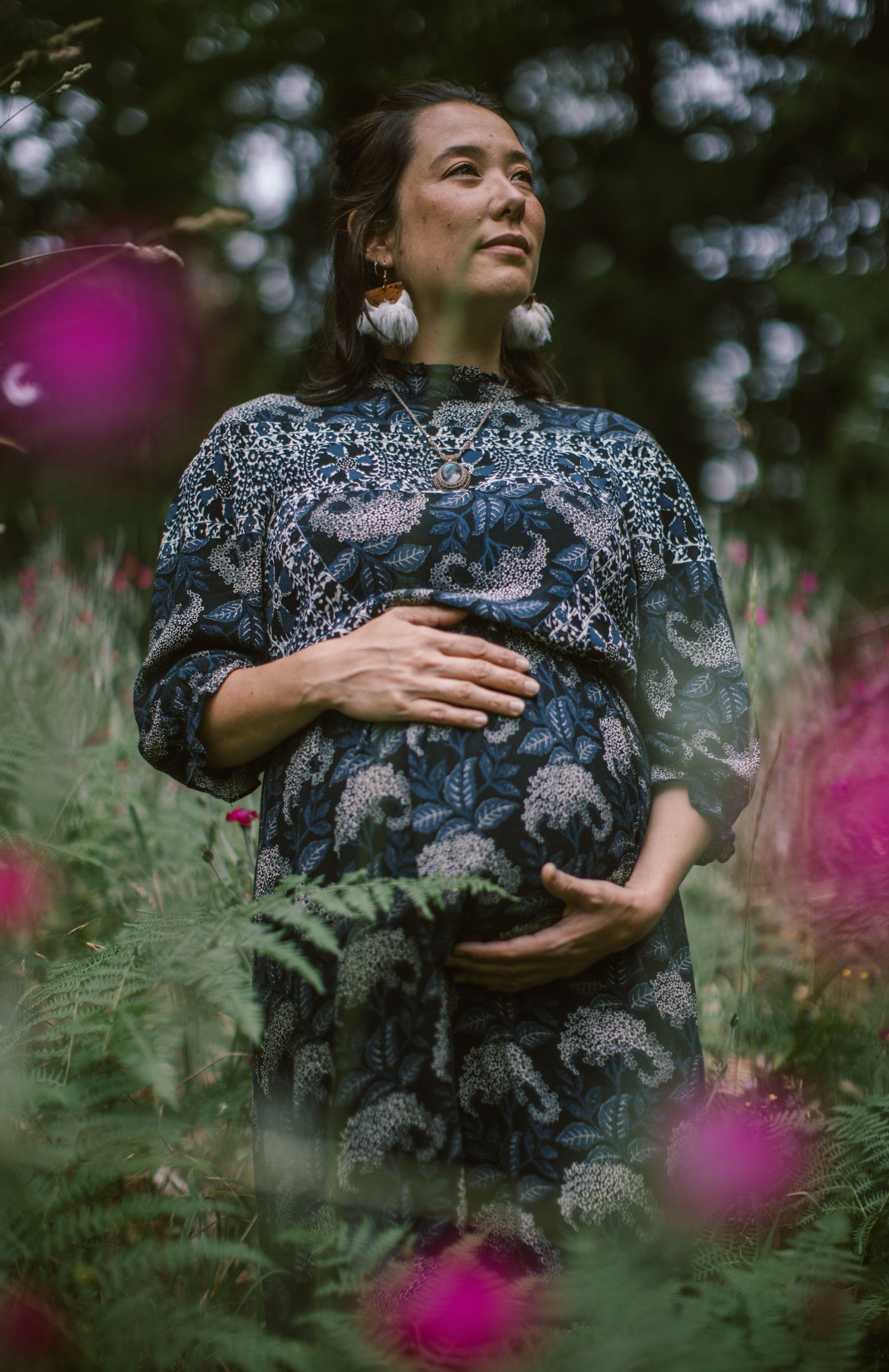 Pregnant woman in patterned dress cradling belly among wild pink flowers and green foliage, gazing upward in serene woodland setting