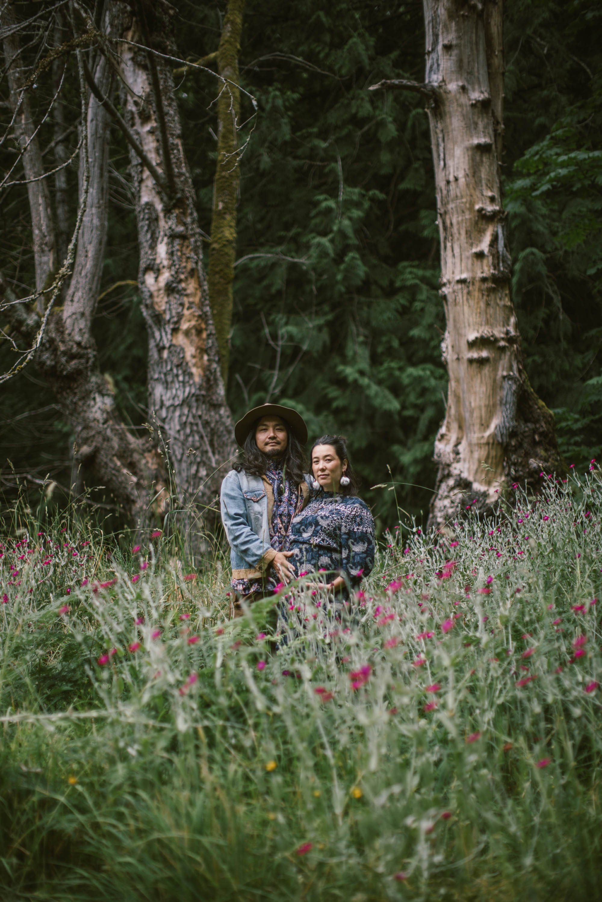 Expectant couple standing among wildflowers between tall evergreen trees in dappled forest light