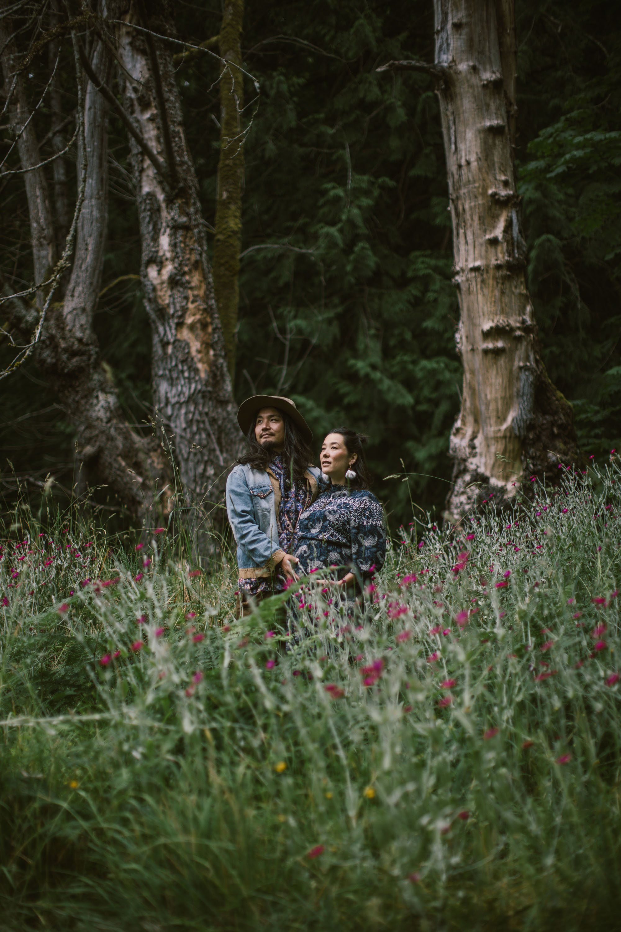 Expectant couple standing among wildflowers in a forest clearing, dappled light filtering through tall birch trees