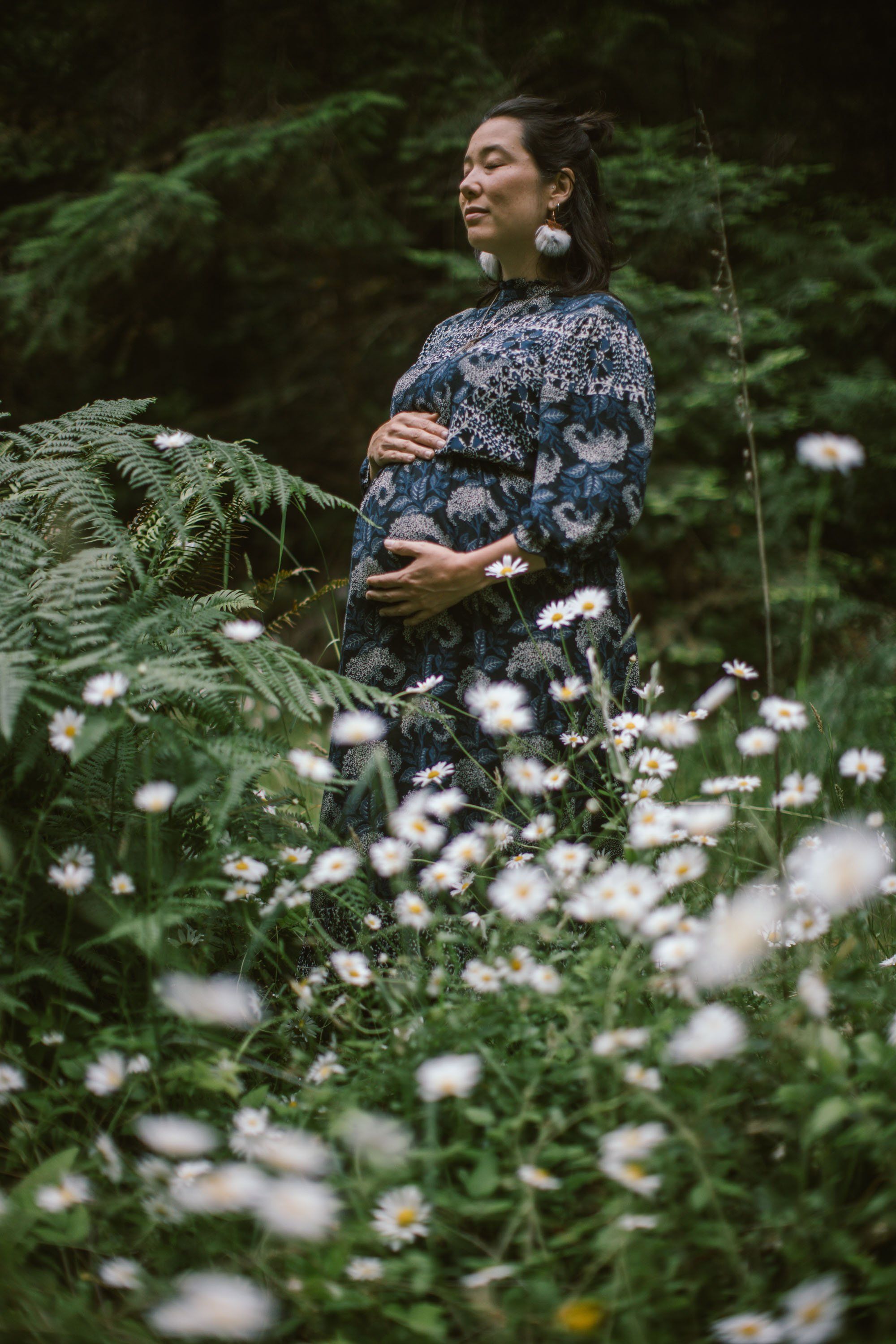 Pregnant woman in patterned blue dress stands among white wildflowers and ferns, gazing thoughtfully to the side in a lush forest setting